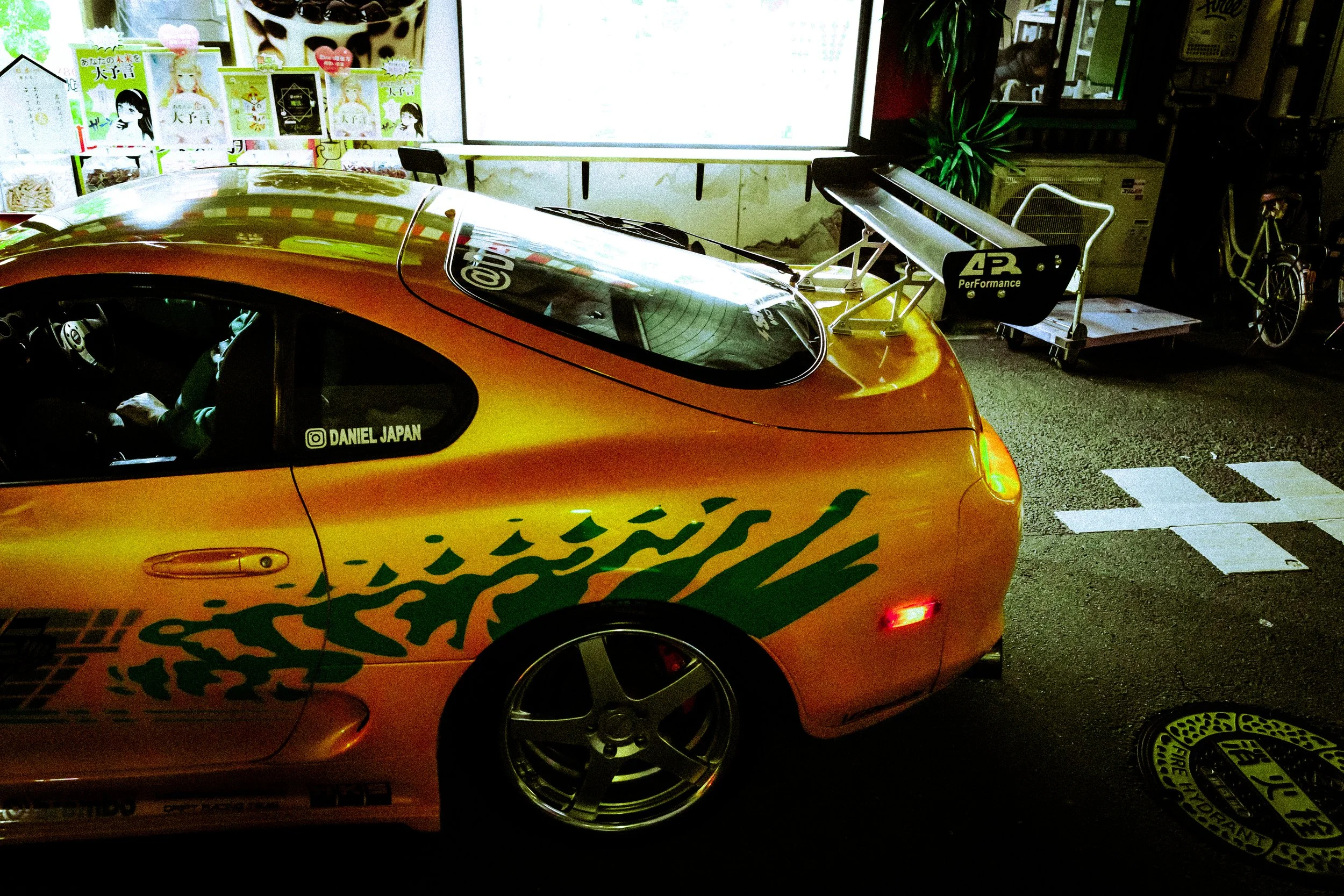 A yellow race car with orange and green decals parked indoors. The window shows a driver sitting inside, and the side of the car displays the Instagram handle '@ DANIEL JAPAN'. There are posters on the wall with Japanese text and drawings, and a raci