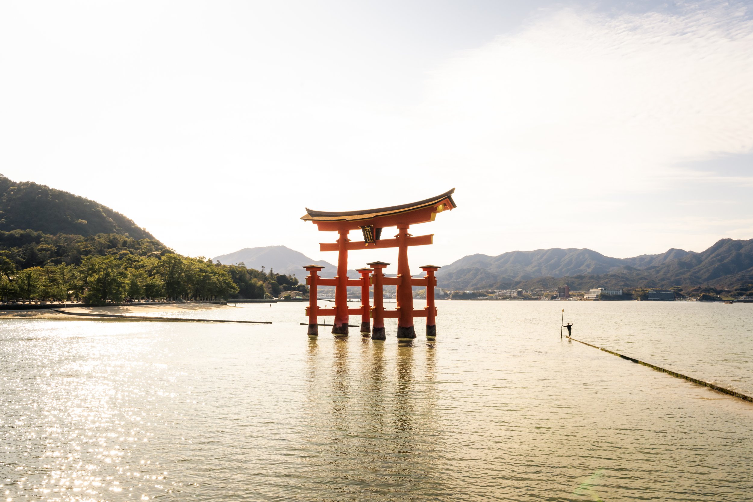 A traditional red Japanese torii gate standing in the water near the shore, with mountains in the background and a person in the water holding a pole near the right side of the image.