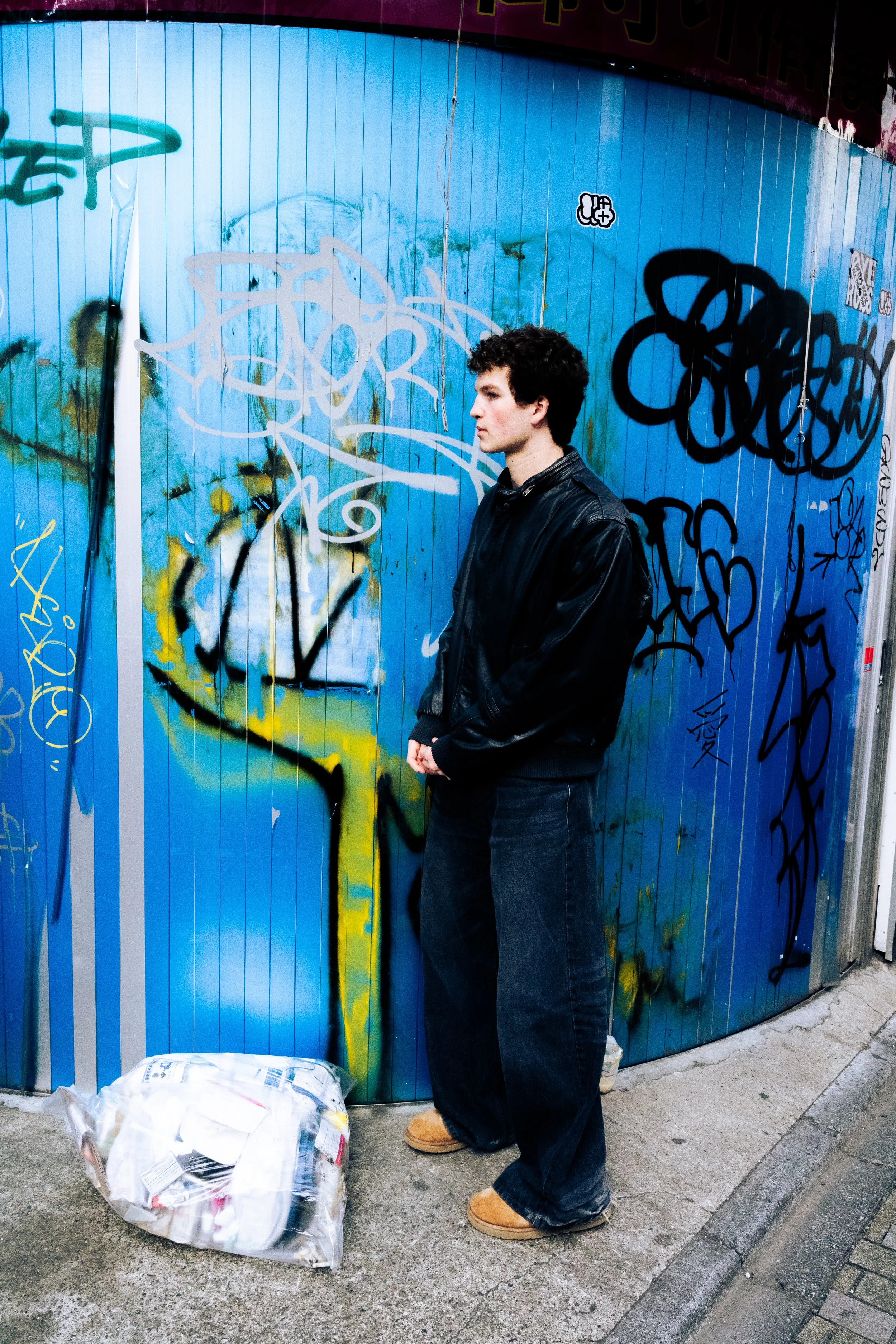 A young man with curly hair, wearing a black leather jacket, dark jeans, and tan boots, stands on a sidewalk with his hands clasped in front of him. He is in front of a blue metal wall with colorful graffiti and stickers. A white plastic bag filled with items is on the ground nearby.