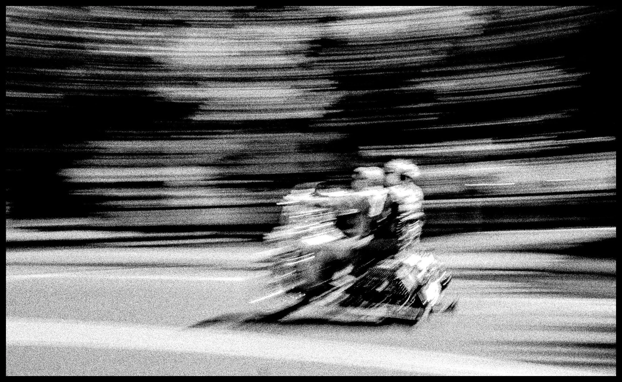 A black and white photograph of two people riding a motorcycle at night. The motion blur gives a sense of speed and movement.