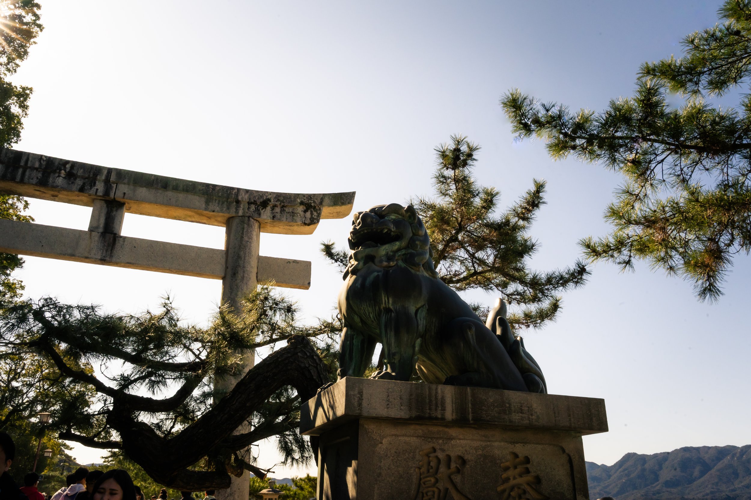A traditional Japanese torii gate with a dragon statue in the foreground, set among trees with mountains and a clear sky in the background.