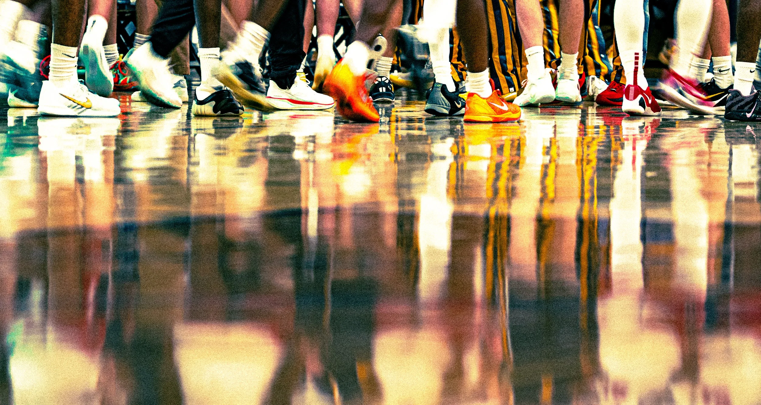 Close-up of people’s legs and feet wearing sports shoes, standing on a shiny wooden floor, likely during a basketball game or practice.