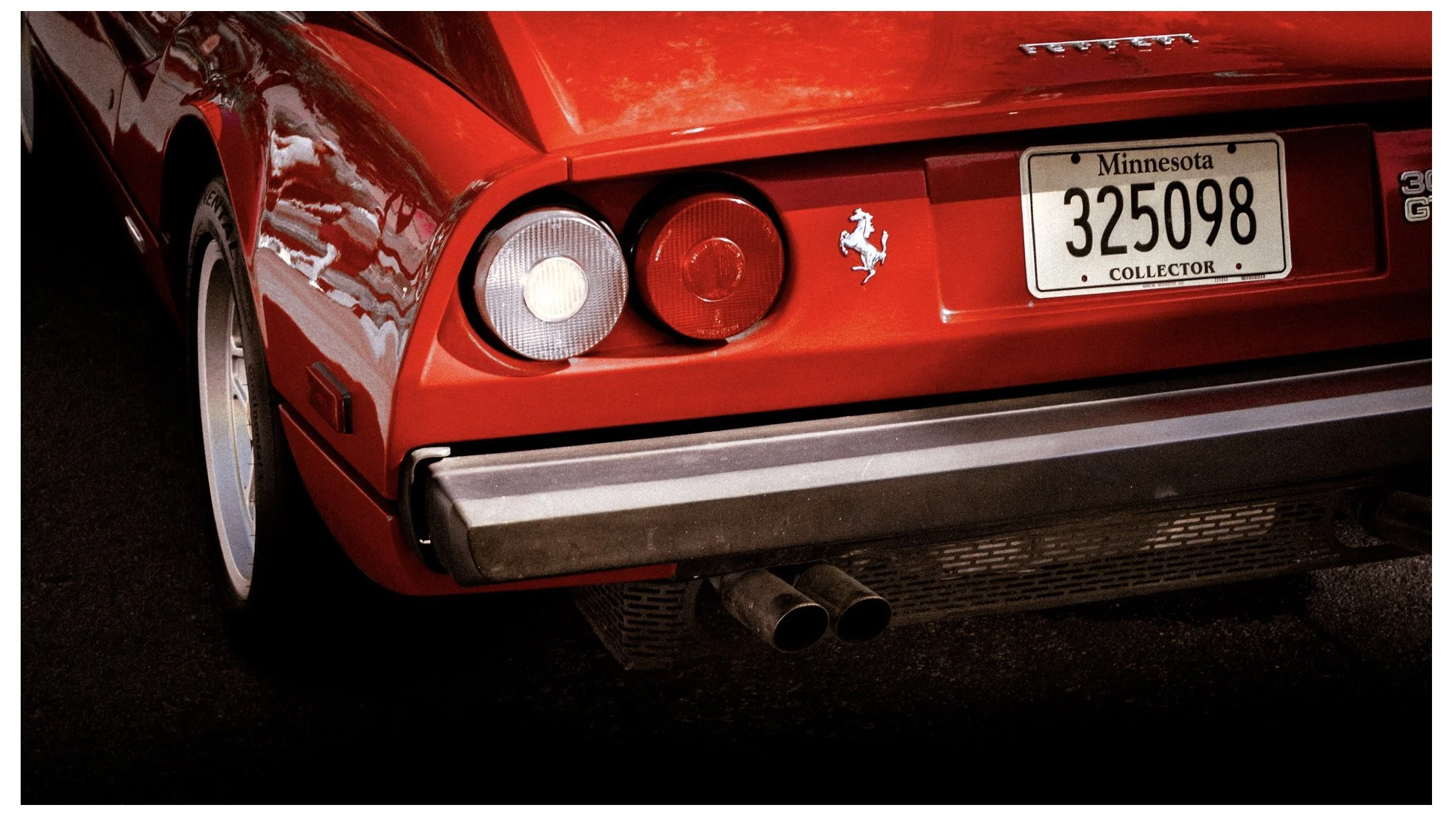 Close-up of the rear of a red Ferrari sports car, showing the taillights, a prancing horse emblem, Minnesota collector license plate, and dual exhaust pipes.