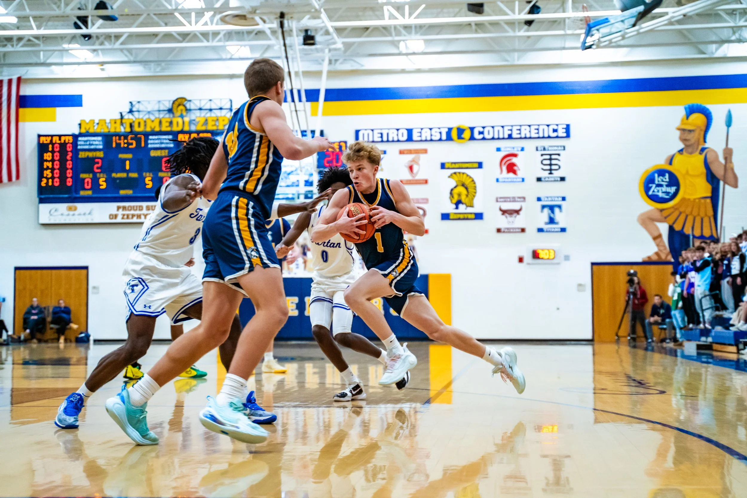 Young basketball players in blue and white uniforms compete during a game in a gymnasium with a large scoreboard and a Spartan mascot statue in the background.