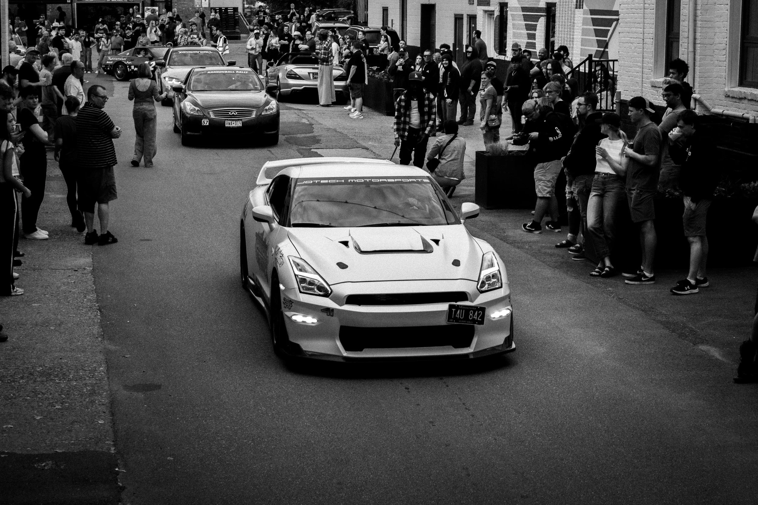 Street scene with a white sports car in the foreground, surrounded by onlookers and parked cars, in an urban area with brick buildings and a crowd gathered on the sidewalk.