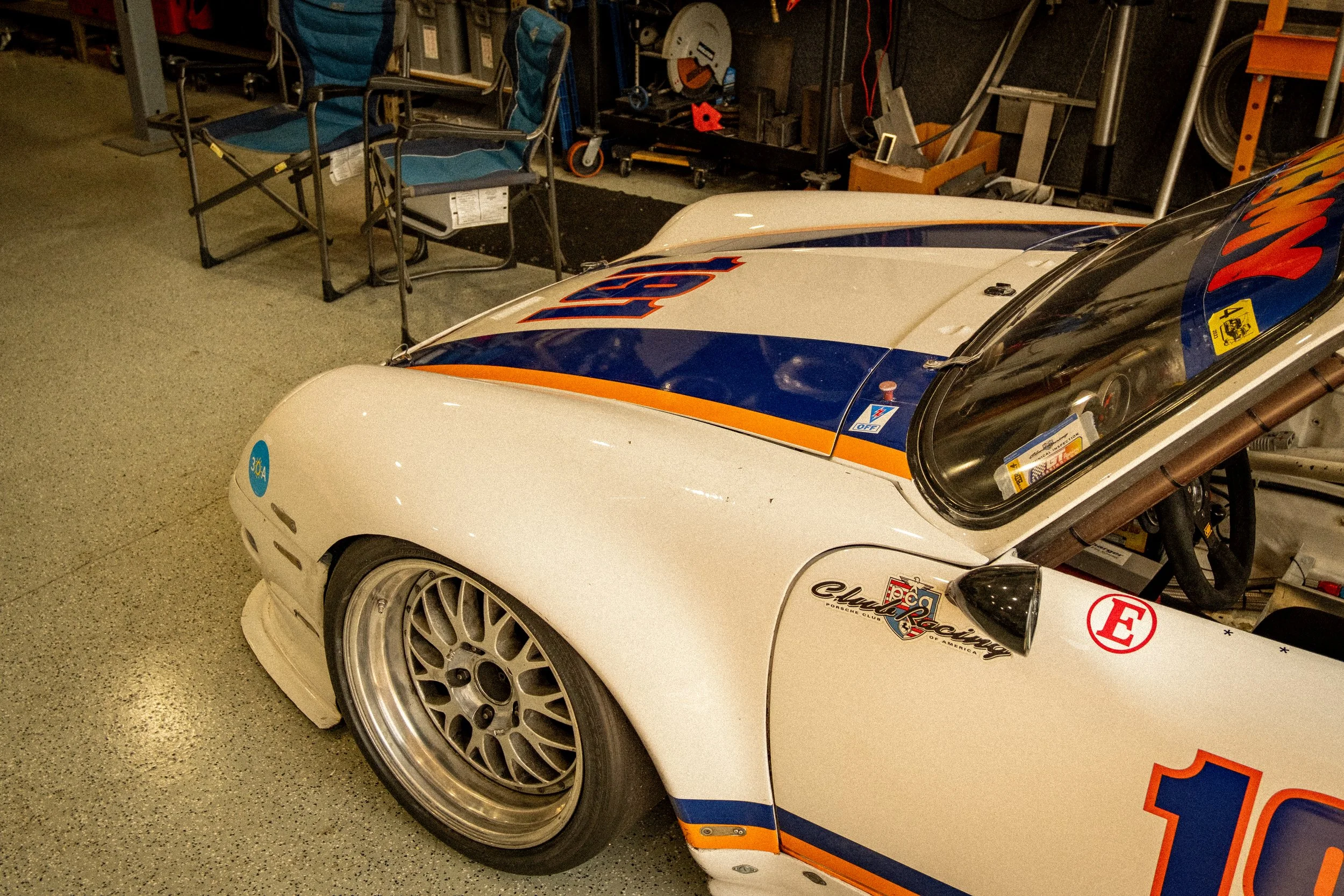 Part of a race car with racing decals, a white front bumper, and large silver alloy wheels, parked in a cluttered garage with chairs and tools in the background.
