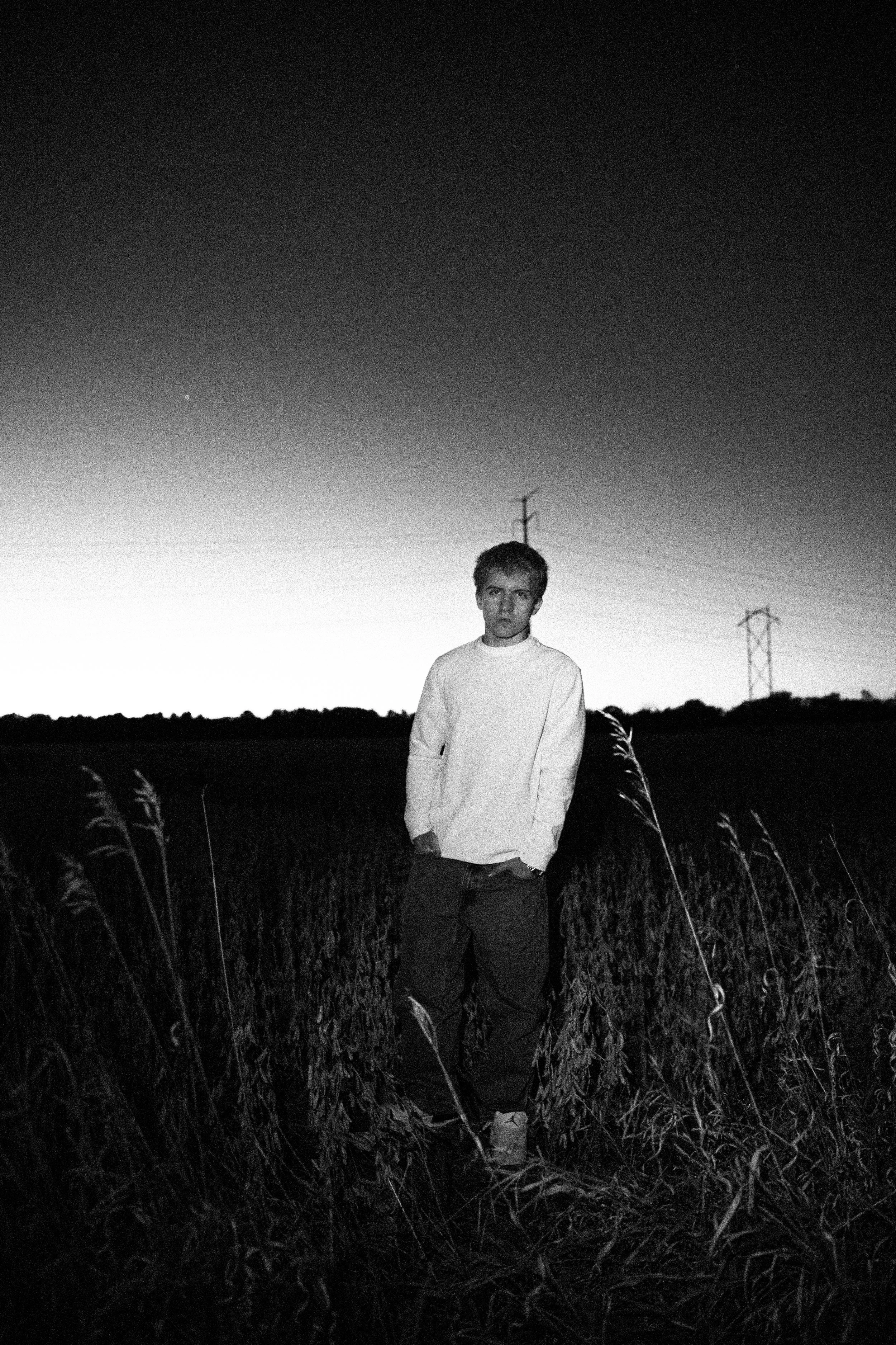 A young man standing in a field at dusk with tall plants and power lines in the background, black and white photo.