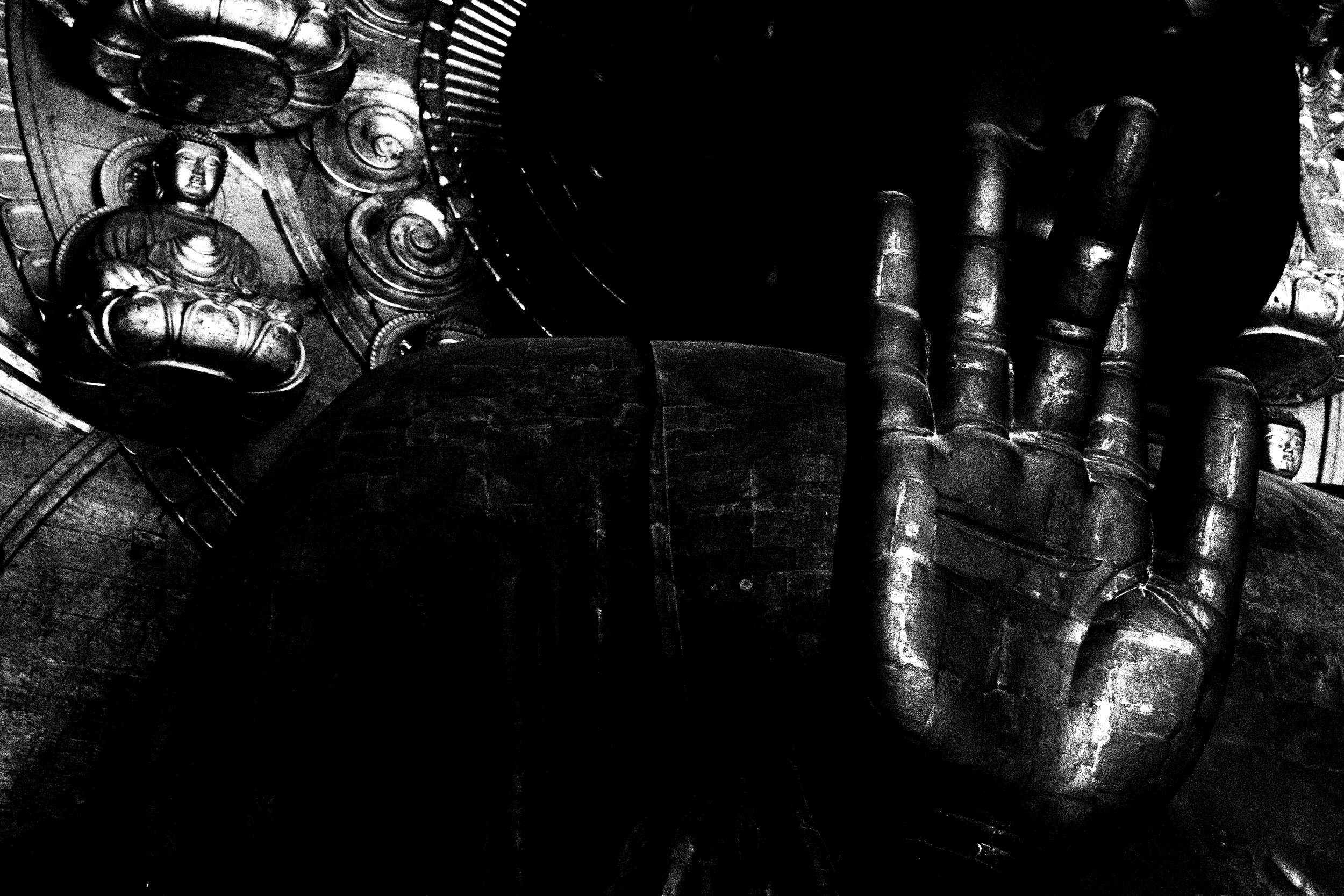 Black and white image of a person wearing dark gloves, holding a bunch of coins and a small Buddha statuette, in a cluttered environment with a metal grate and other objects.