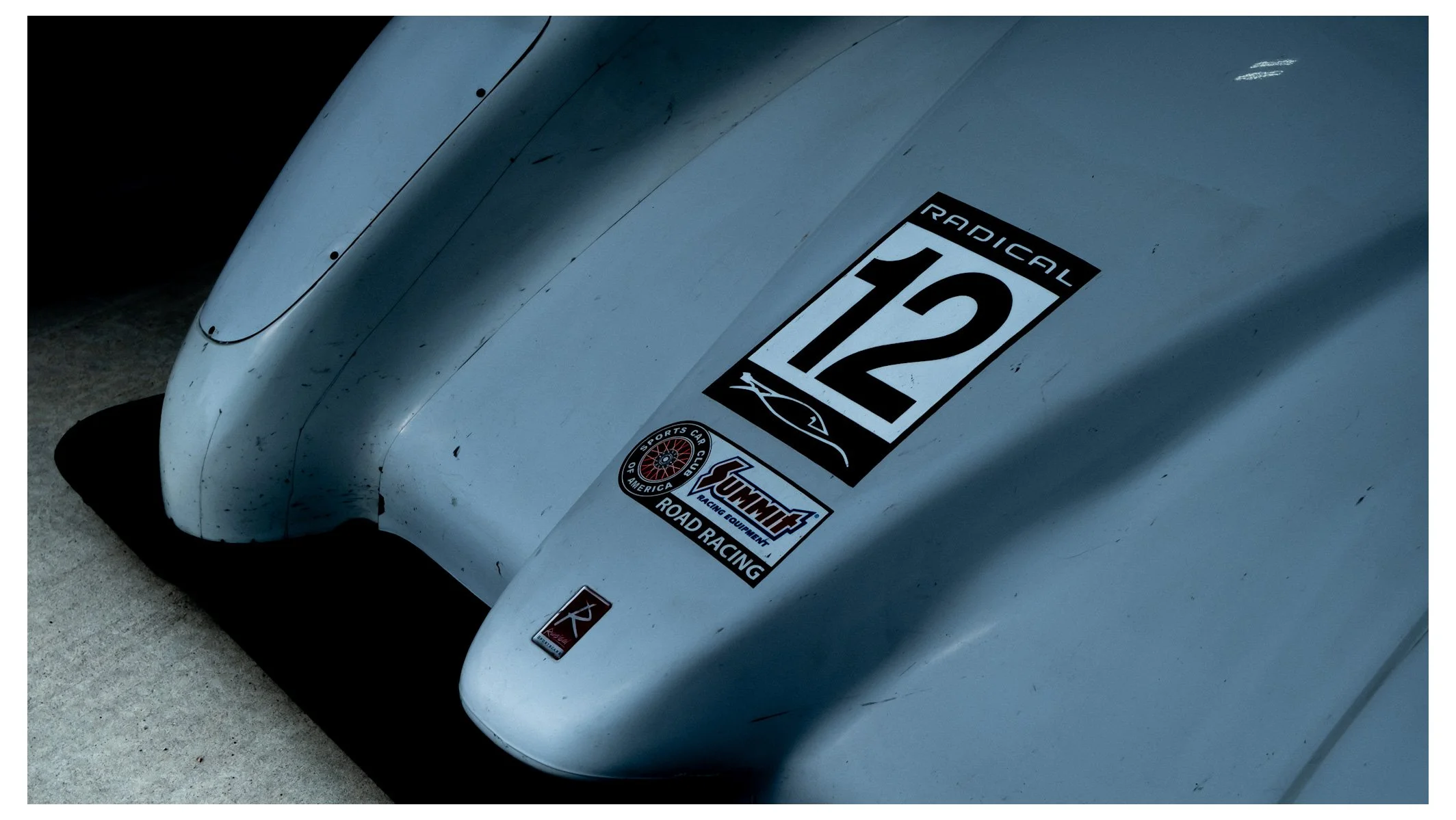Close-up of the front of a race car, featuring a sticker with the number 12, and various racing-related logos. The car is silver with visible dirt and scratches.