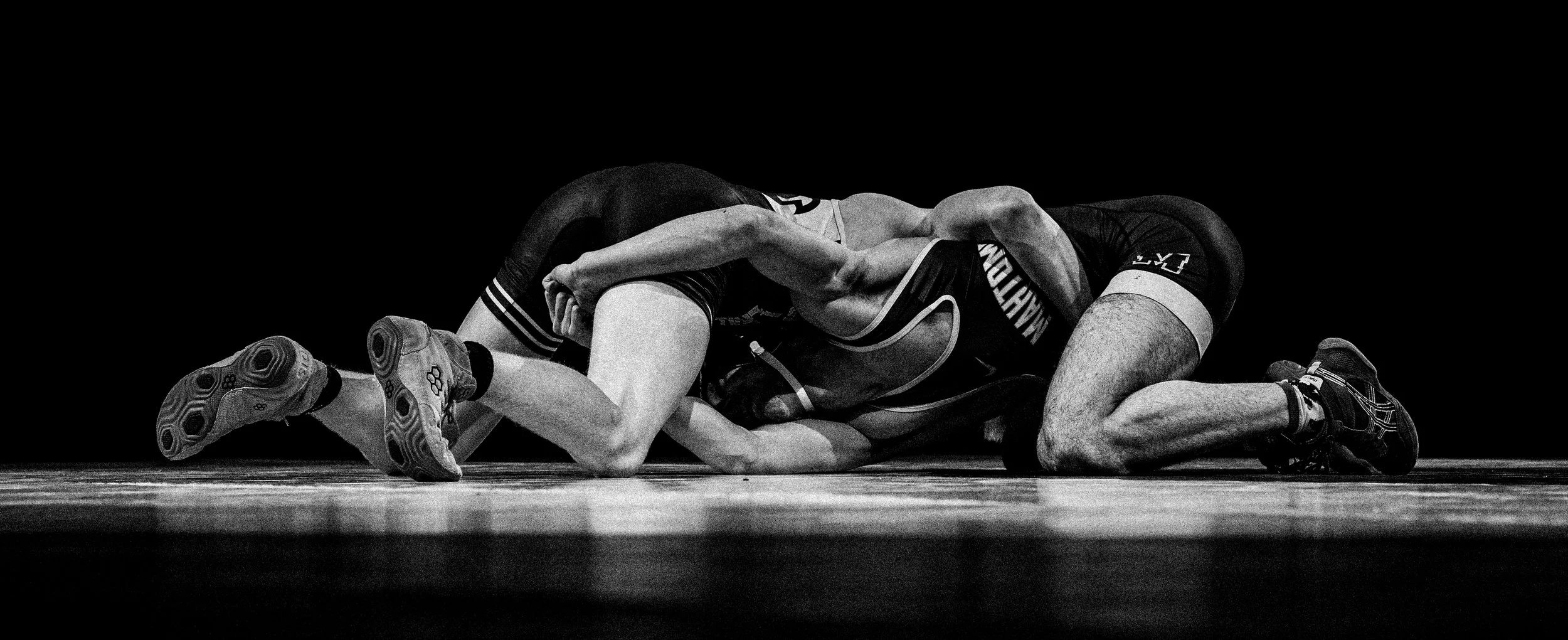 Two wrestlers on the mat during a wrestling match, one in black and one in light-colored uniforms, in a black and white photo against a dark background.