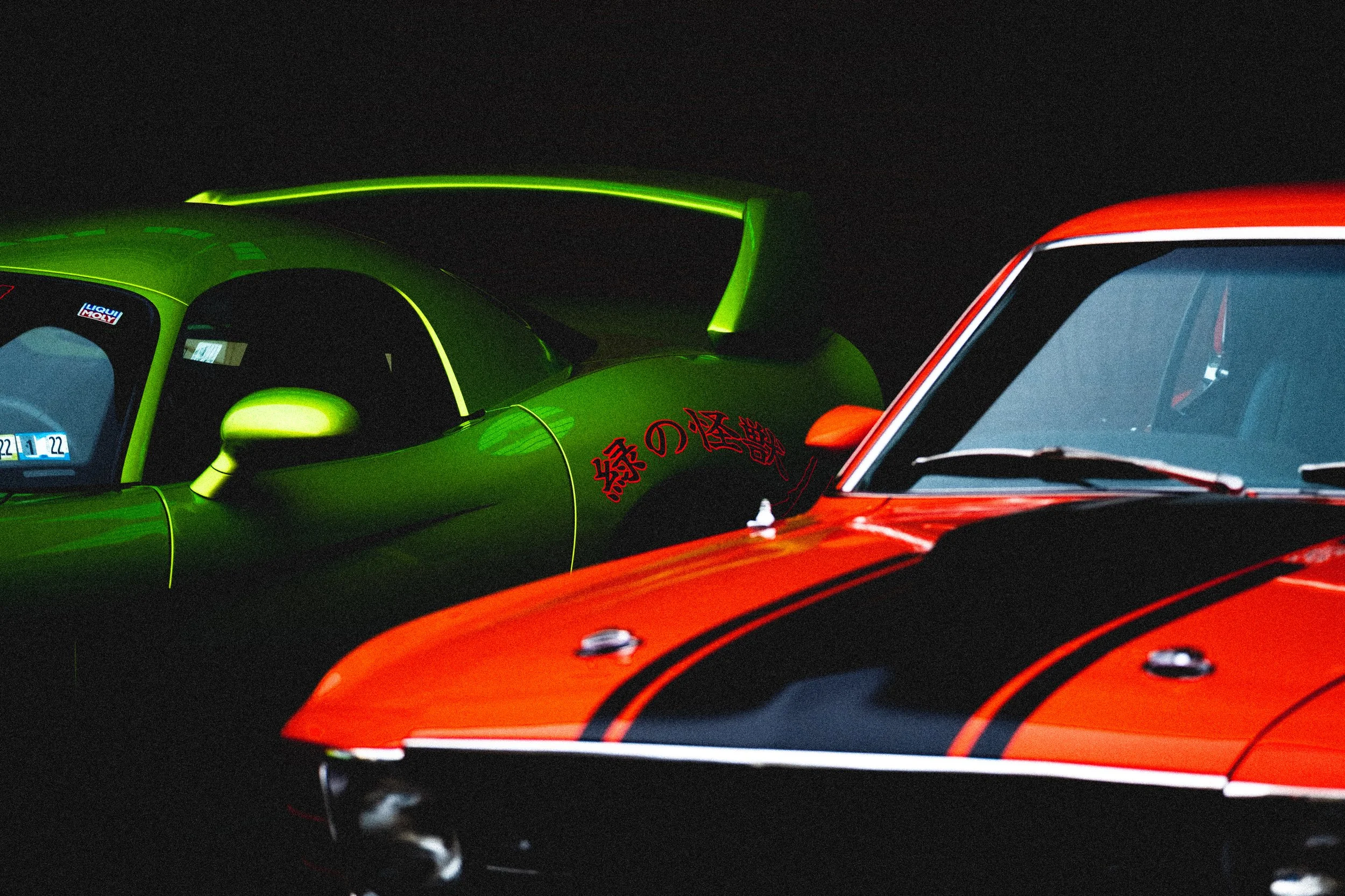 Close-up of two sports cars, one green and one red, parked side by side at night. The green car has Japanese characters on the side and a large rear wing, while the red car features black stripes and a visible side mirror.
