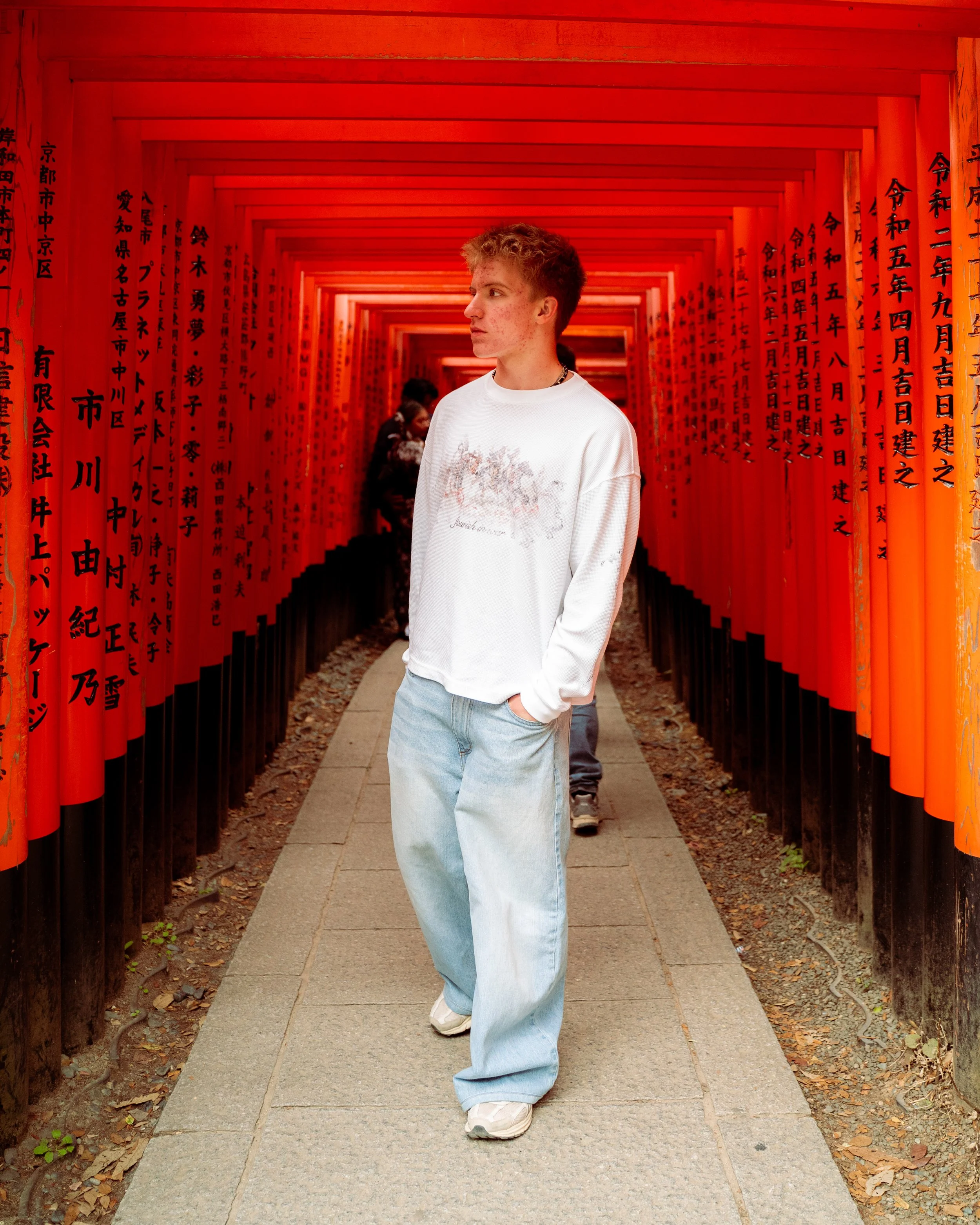 A young man with curly hair and light skin stands in front of bright red torii gates with black Japanese inscriptions in a pathway at a Japanese shrine. There are other visitors behind him.