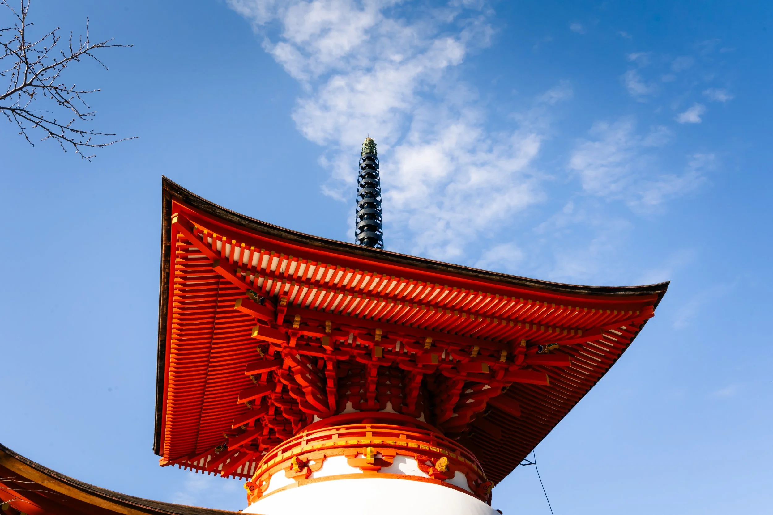 Close-up view of the top part of a traditional Japanese pagoda with a red-orange roof, a decorative black finial, and a blue sky with scattered clouds in the background.