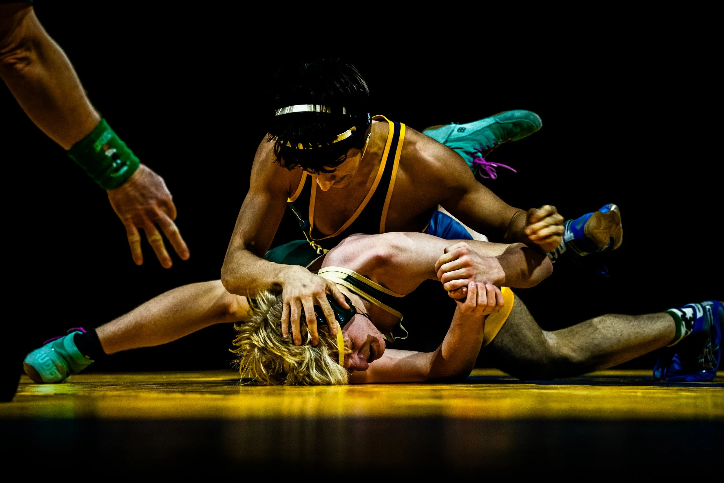 Two wrestlers on the mat; one wrestler is on top, applying a hold on the other who is lying face down, with another person reaching toward them.