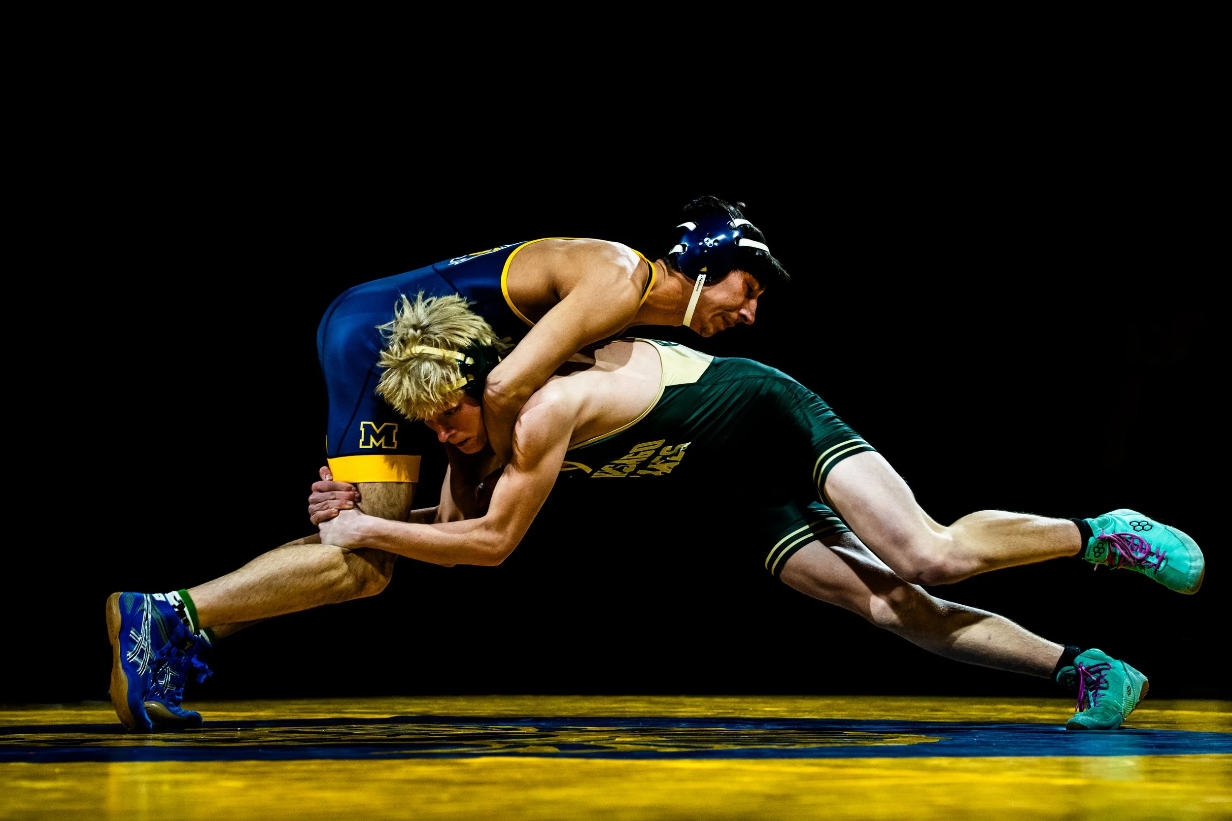 Two male wrestlers competing on a mat, one in a blue singlet with a yellow trim and the other in a green and yellow singlet. The wrestler in blue is on top, pinning the other wrestler.