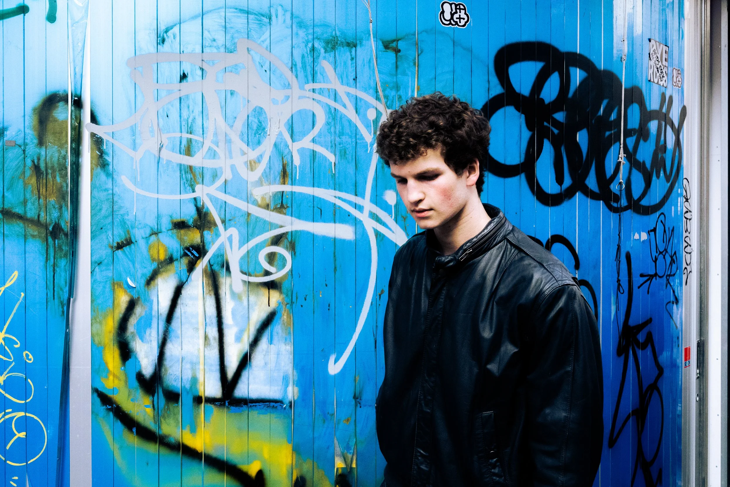 A young man with curly hair and wearing a black leather jacket standing in front of a blue wall covered in various graffiti tags and designs.