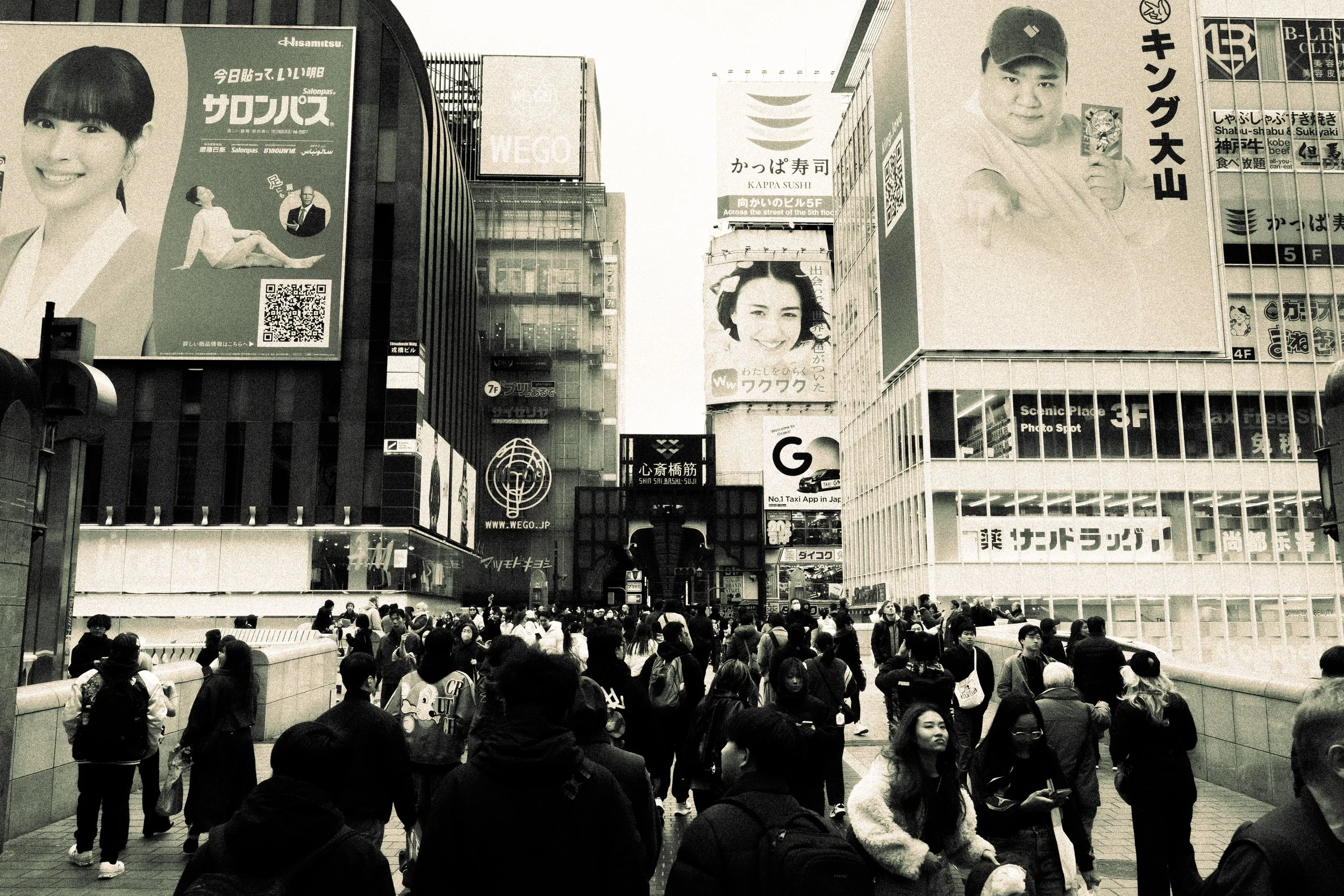 A busy city street scene with many pedestrians. Tall buildings with large advertisements and signs, including a woman smiling, a man in a baseball cap, and various billboards in Japanese and English. The atmosphere is bustling and vibrant.