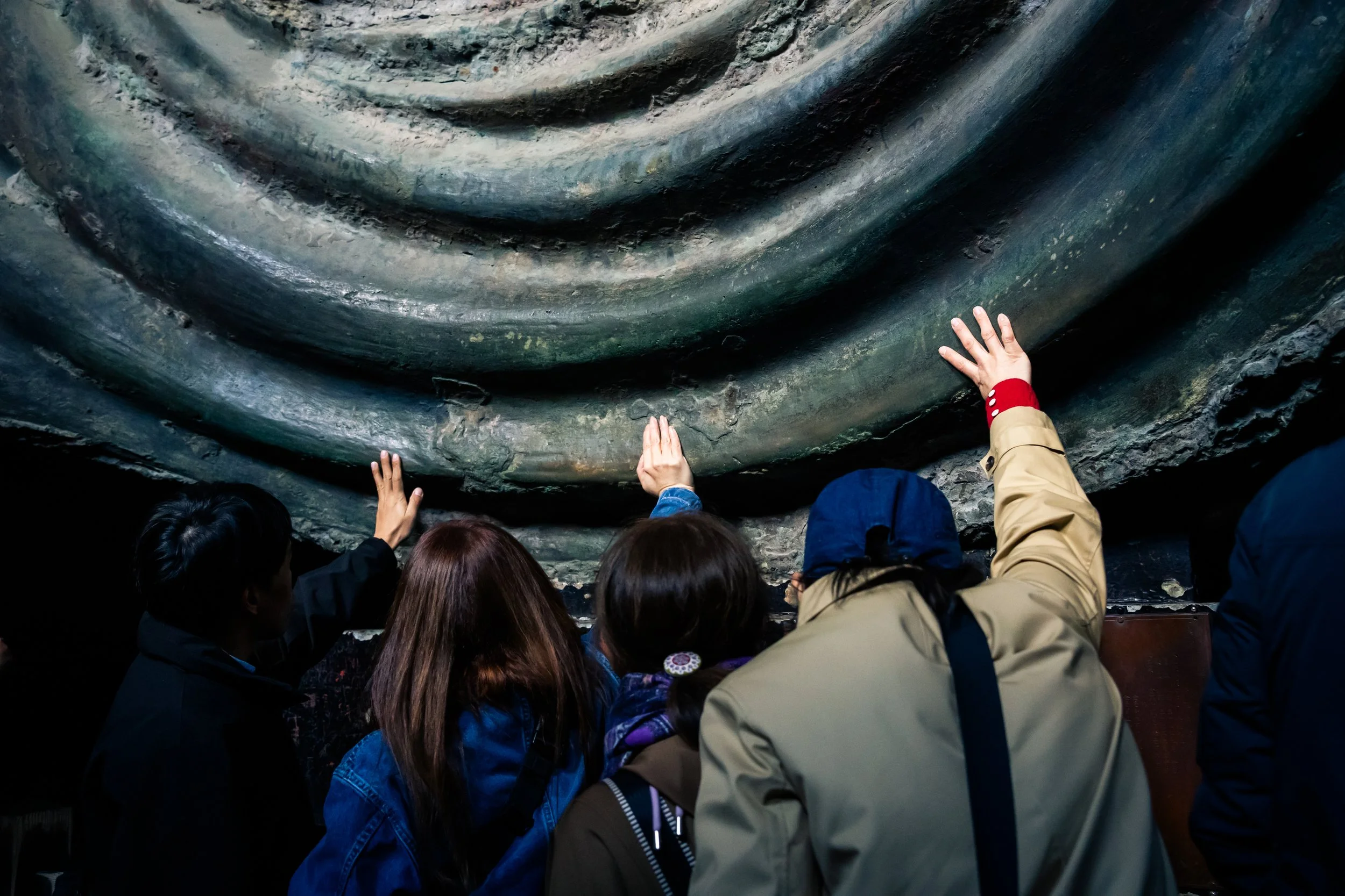 People touching and observing a large, textured, dark-colored geological formation or cave wall.