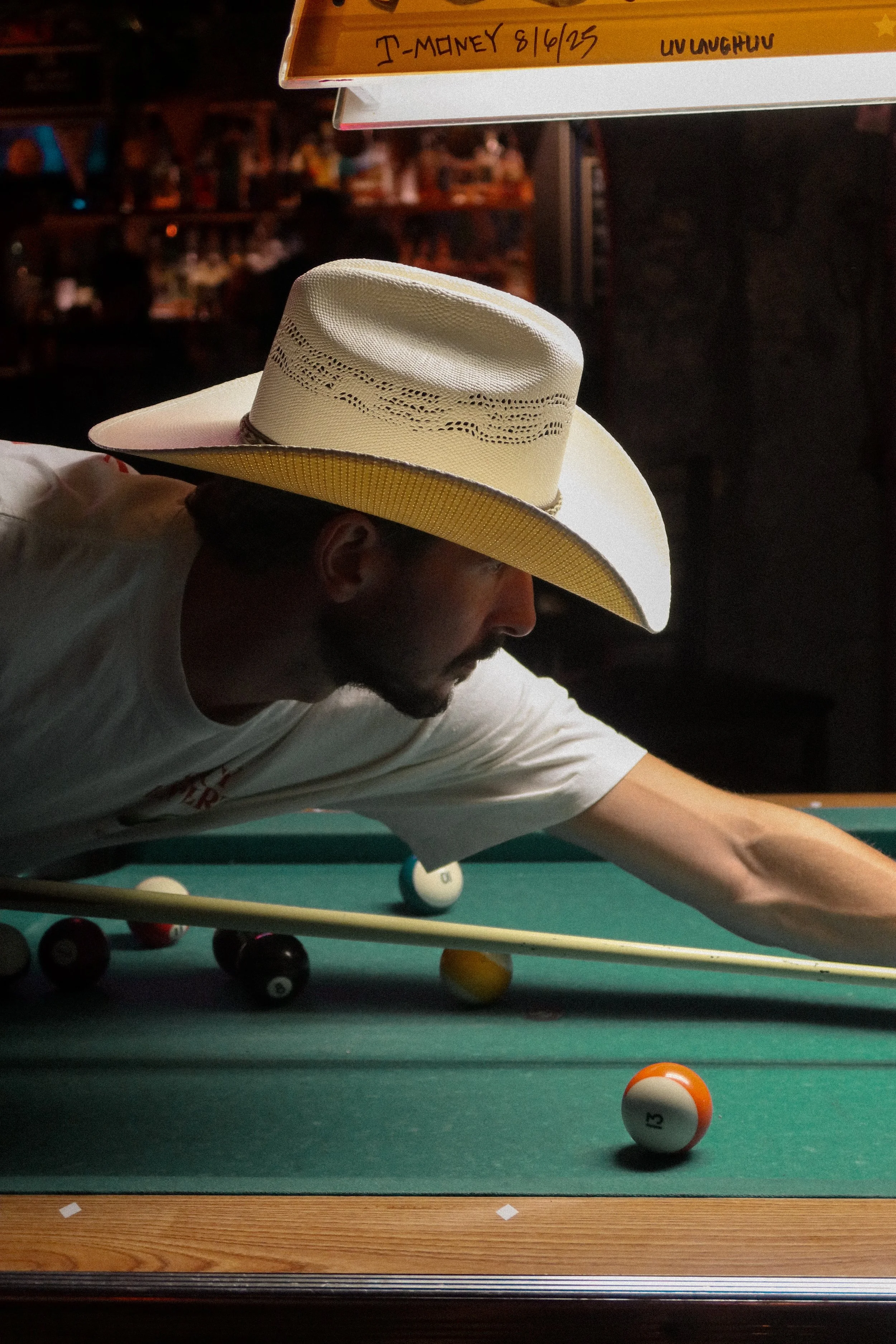 A man in a white shirt wearing a cream-colored cowboy hat playing pool on a green pool table with ball number 15 near the bottom right corner.