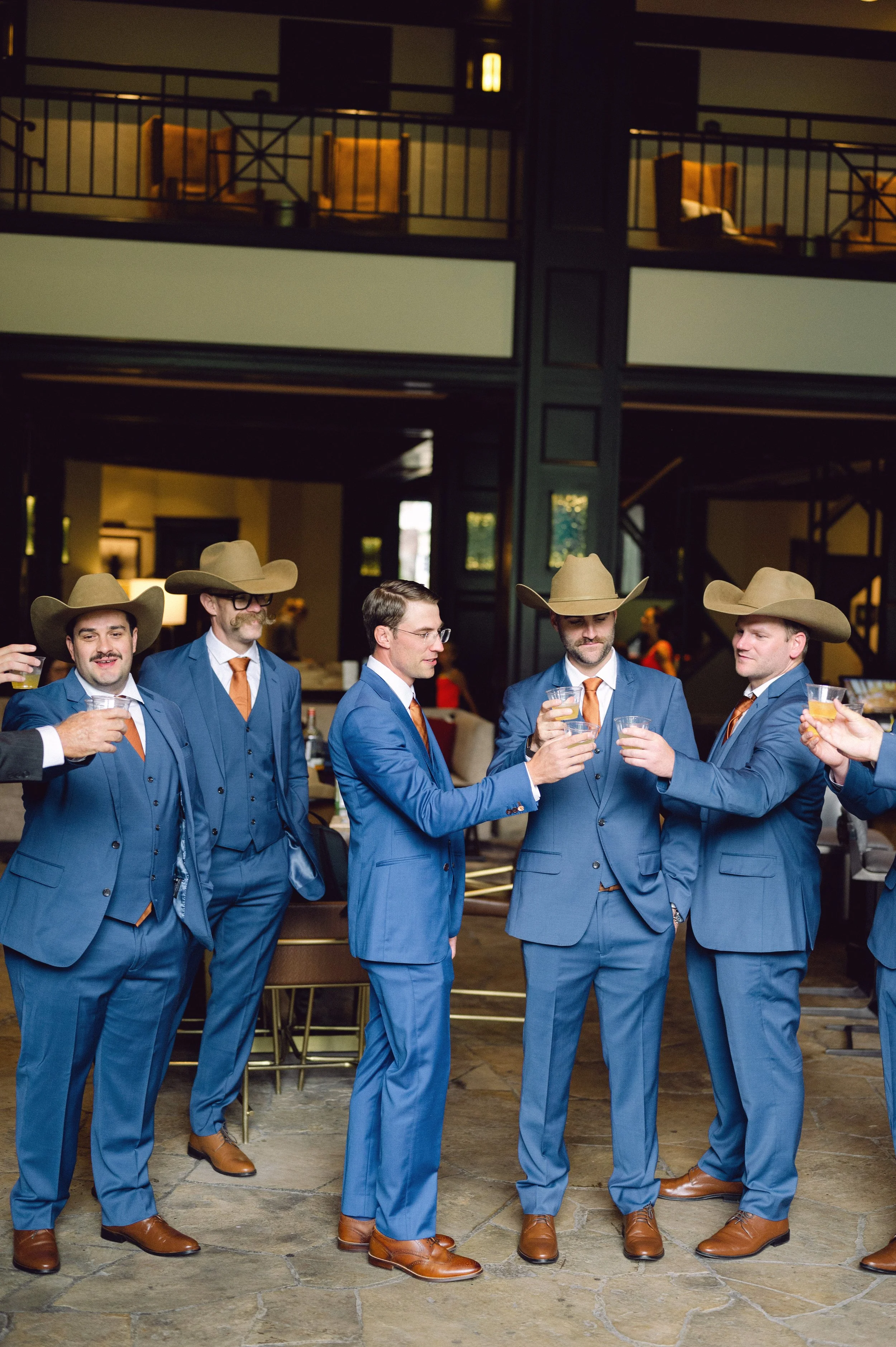 Group of men dressed in blue suits, brown shoes, orange ties, and wearing cowboy hats, standing in a line at a social gathering, holding drinks and engaged in conversation.