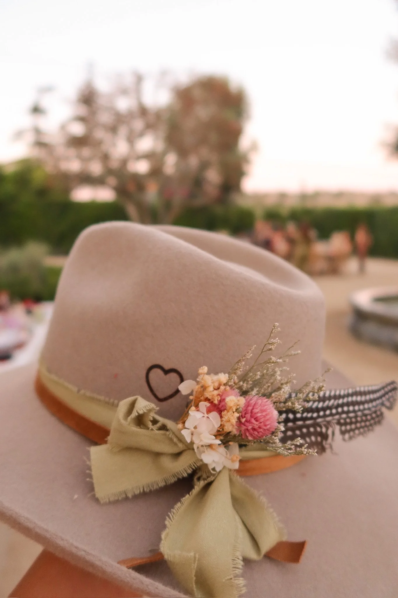 A beige hat decorated with a small bouquet of pink and white flowers and a large green bow, placed on a table with blurred trees and outdoor setting in the background.