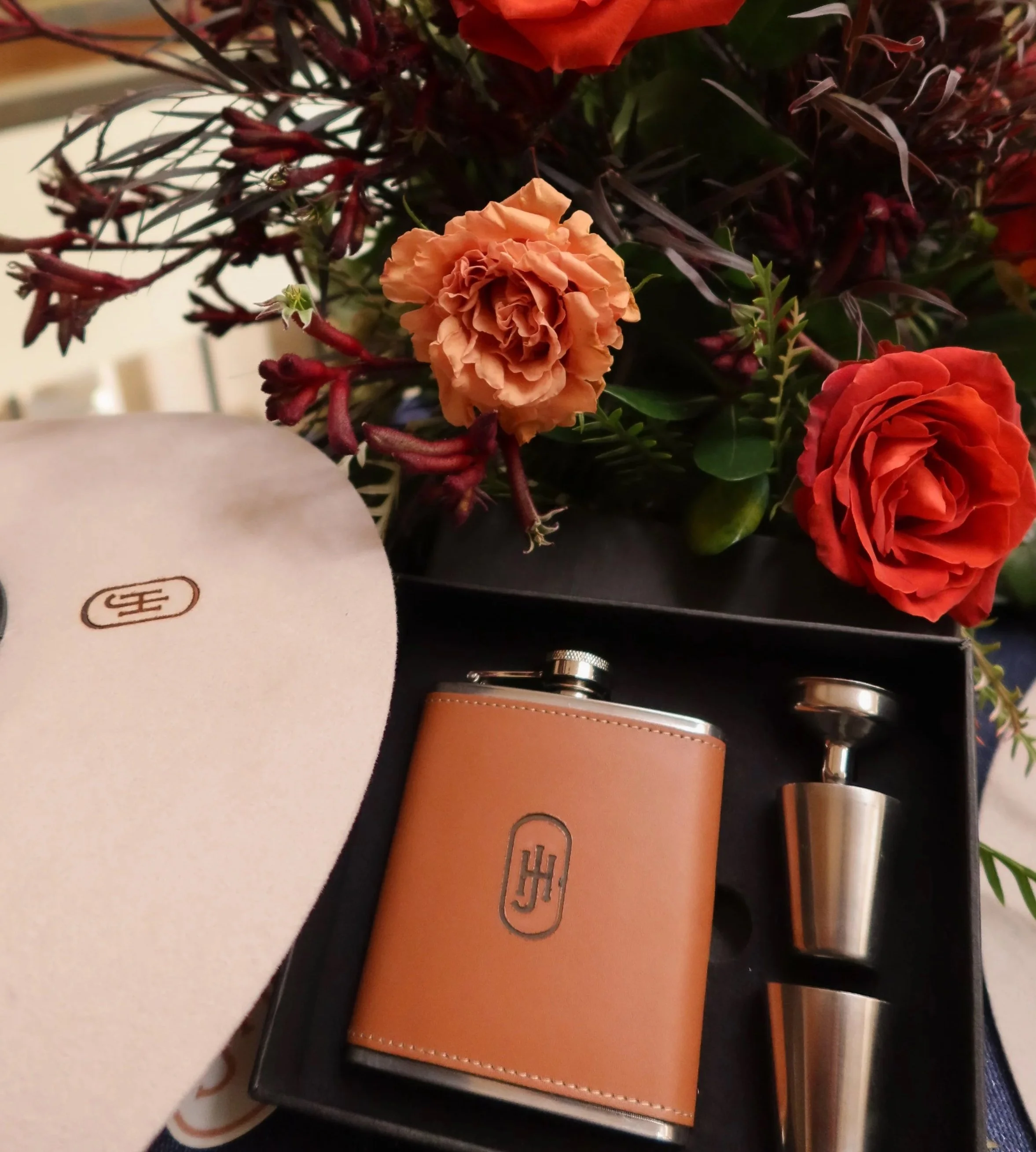 Close-up of a bouquet of orange, red, and pink flowers with green leaves, with a blurred background including a white object, a leather item, and a silver metallic container.