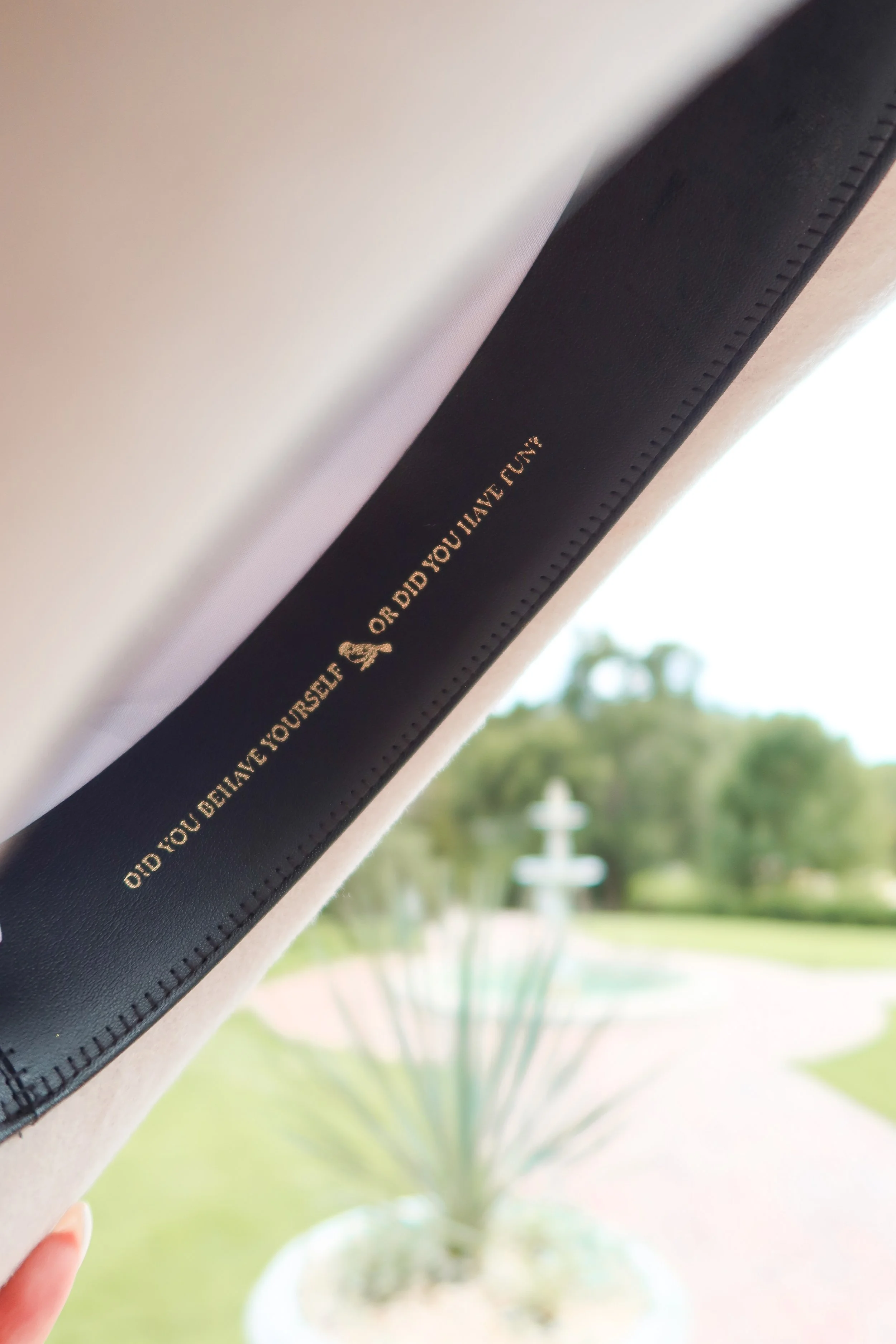 Close-up of a black book or notebook with gold embossed text on the spine, with a blurred green plant and a fountain in the background outside a window.