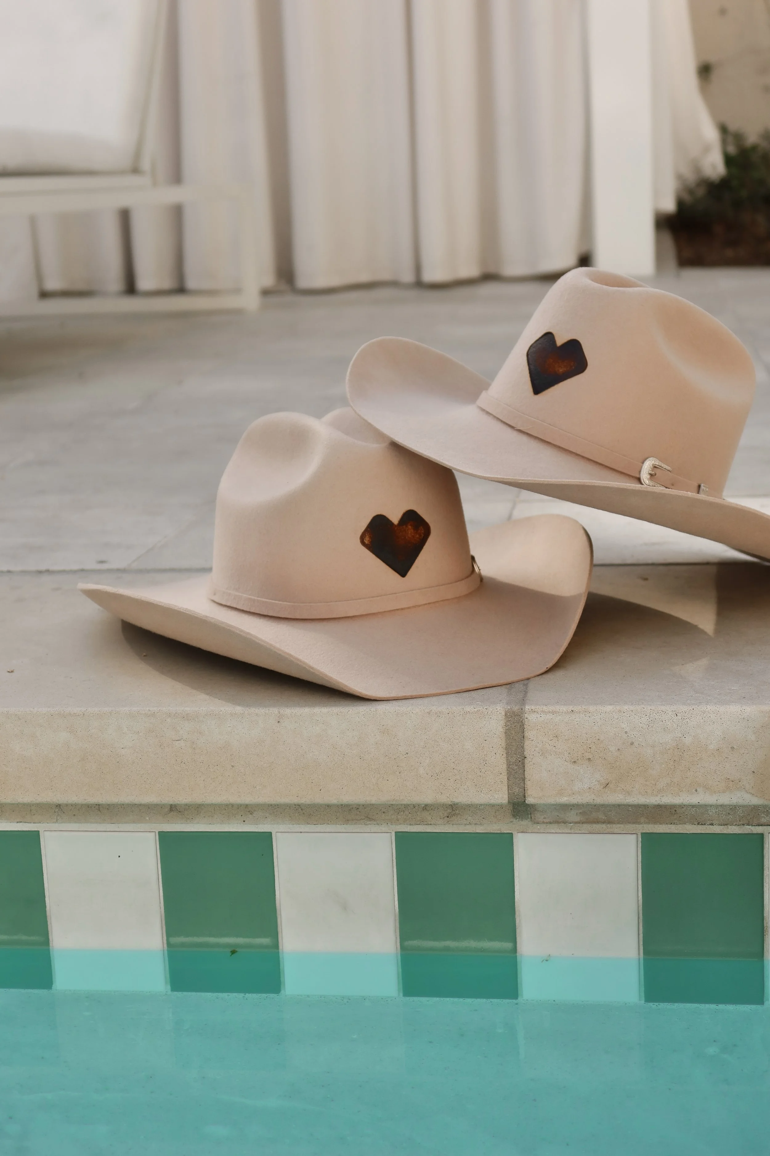 Two beige cowboy hats with black heart-shaped symbols on them resting on a tiled surface, with a partially visible light-colored curtain and greenery in the background.
