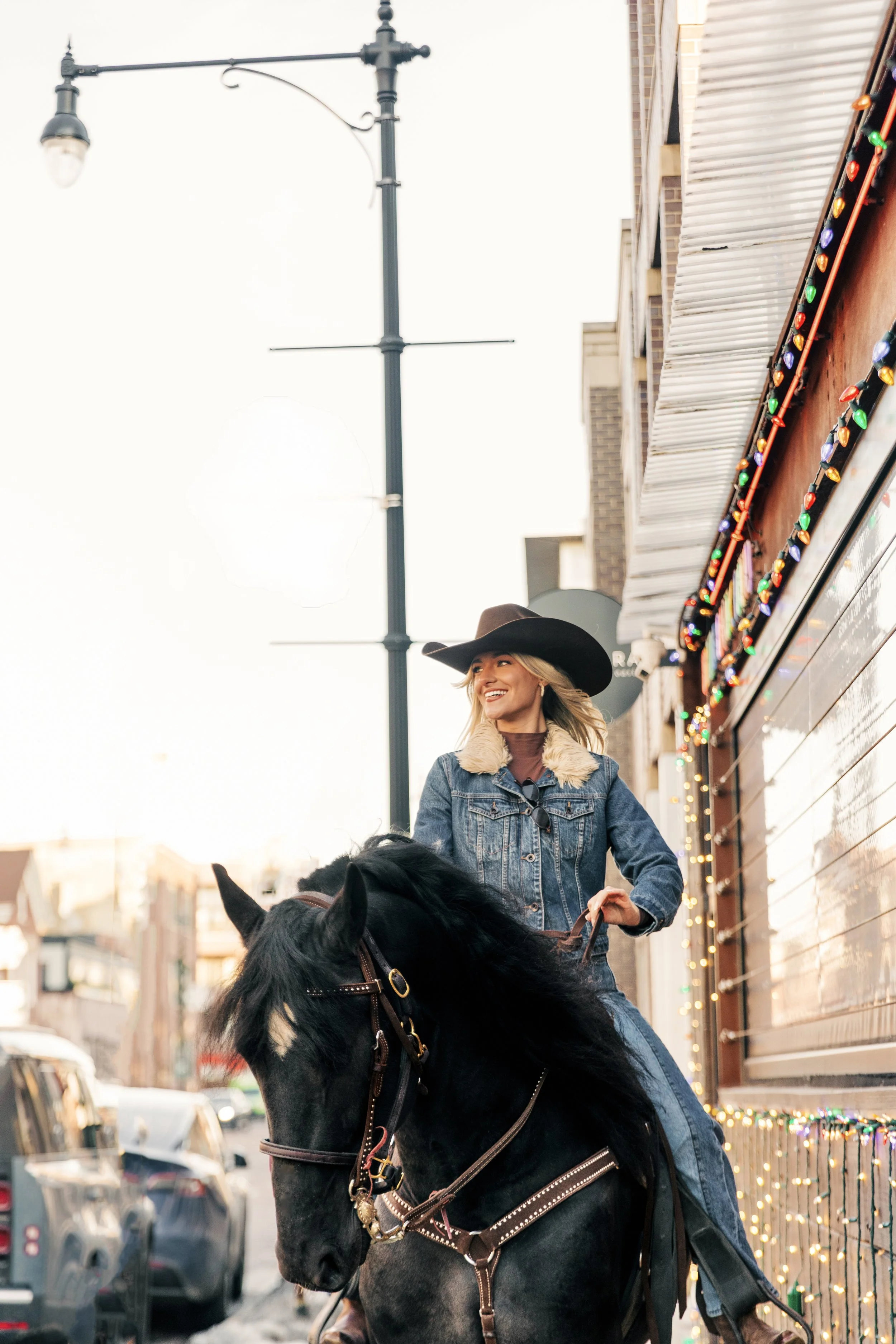 A woman with blonde hair, wearing a black cowboy hat, denim jacket with a fur collar, riding a black horse on a city street with parked cars and a building with colorful string lights and a wooden exterior.