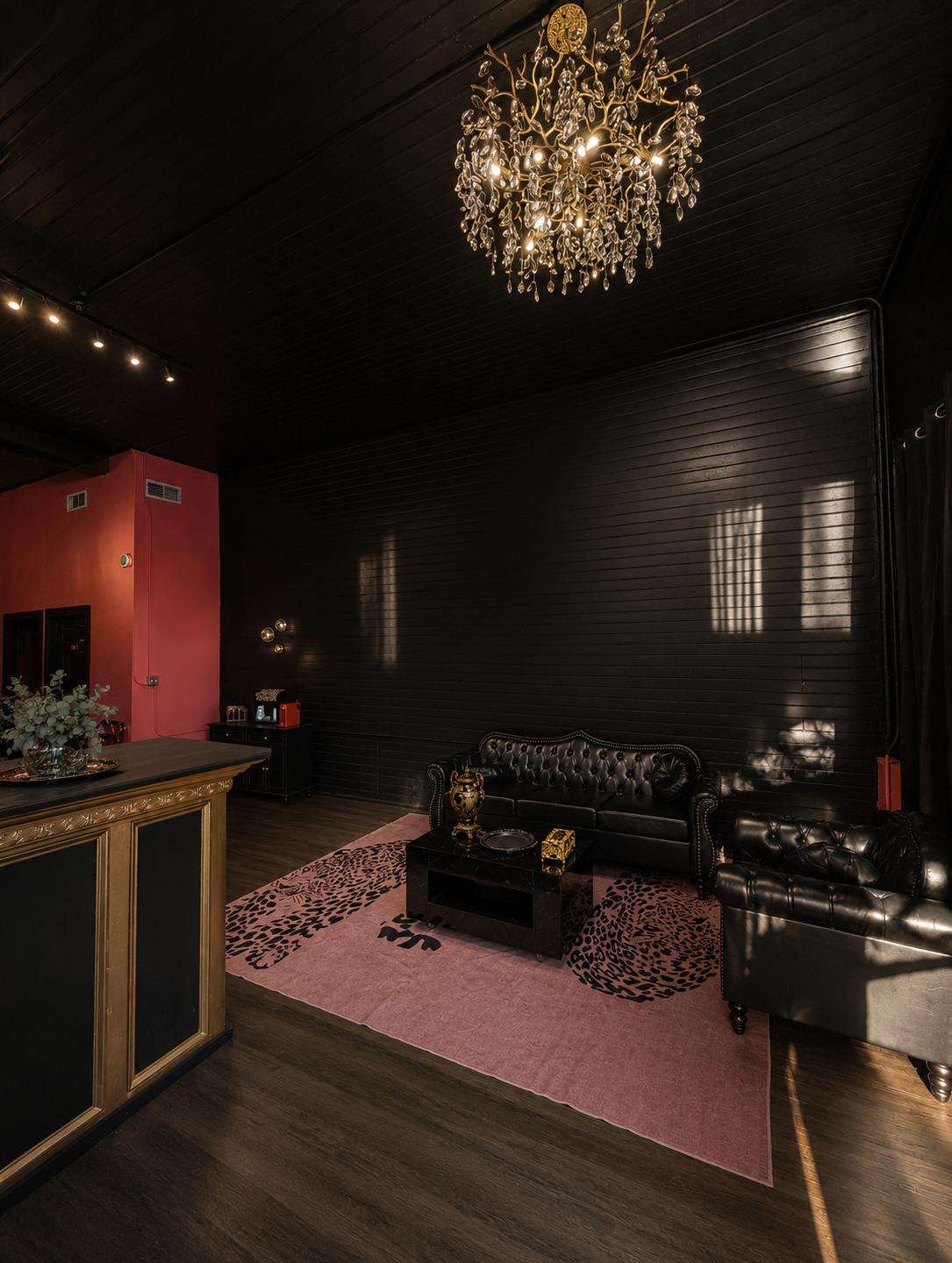A luxurious living room with black leather tufted sofas, a pink rug with black animal print, a black coffee table with decorative items, a gold chandelier, and dark wood flooring and ceiling.