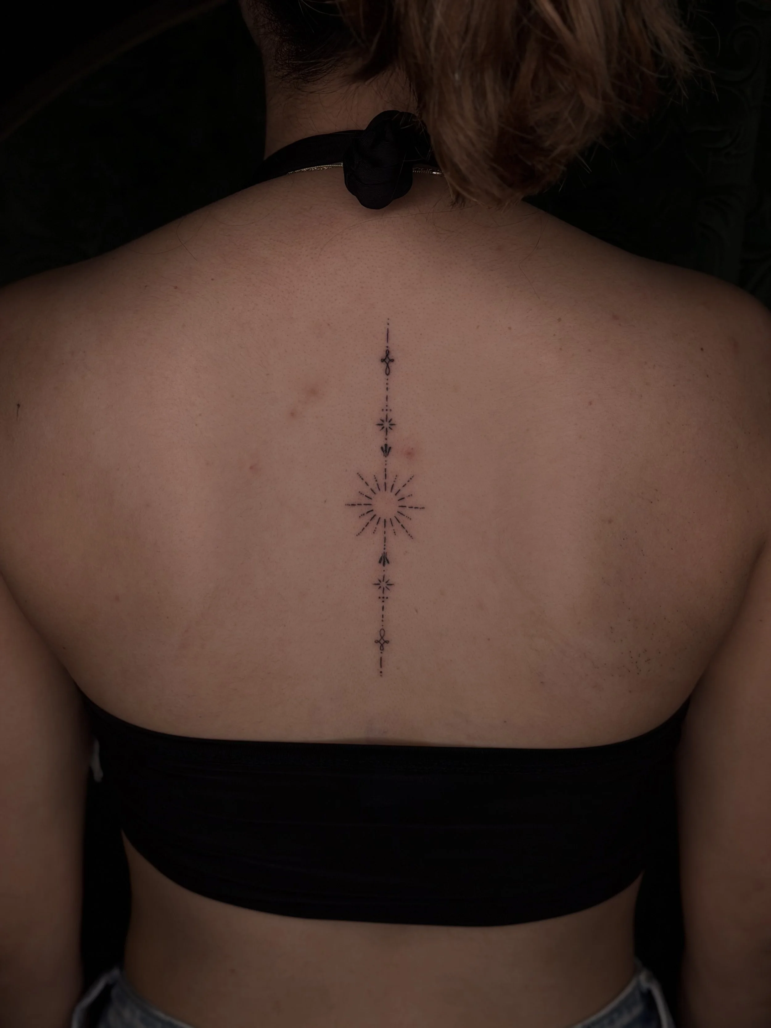 A person's back with a vertical line tattoo running down the center, featuring small decorative elements and sunburst designs, with the person's head slightly bowed and brown hair visible.