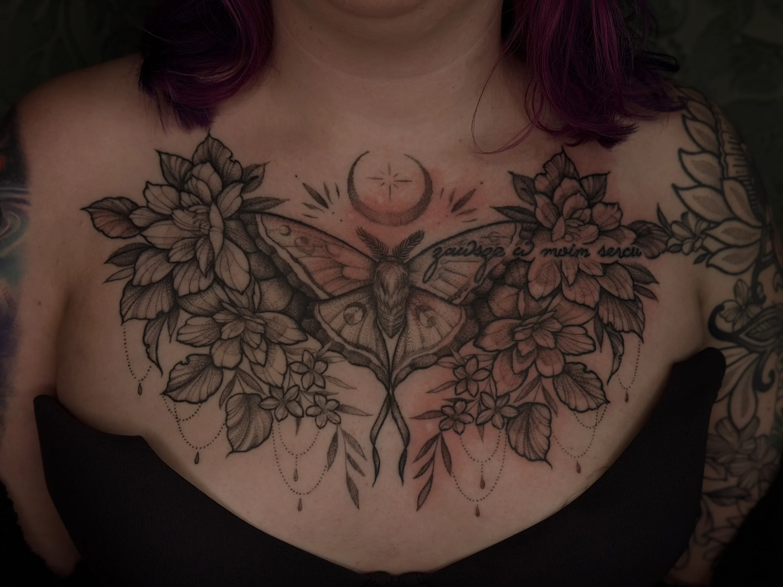 Close-up of a person's chest with a detailed floral tattoo featuring flowers, leaves, a crescent moon, and a butterfly, with some script text.