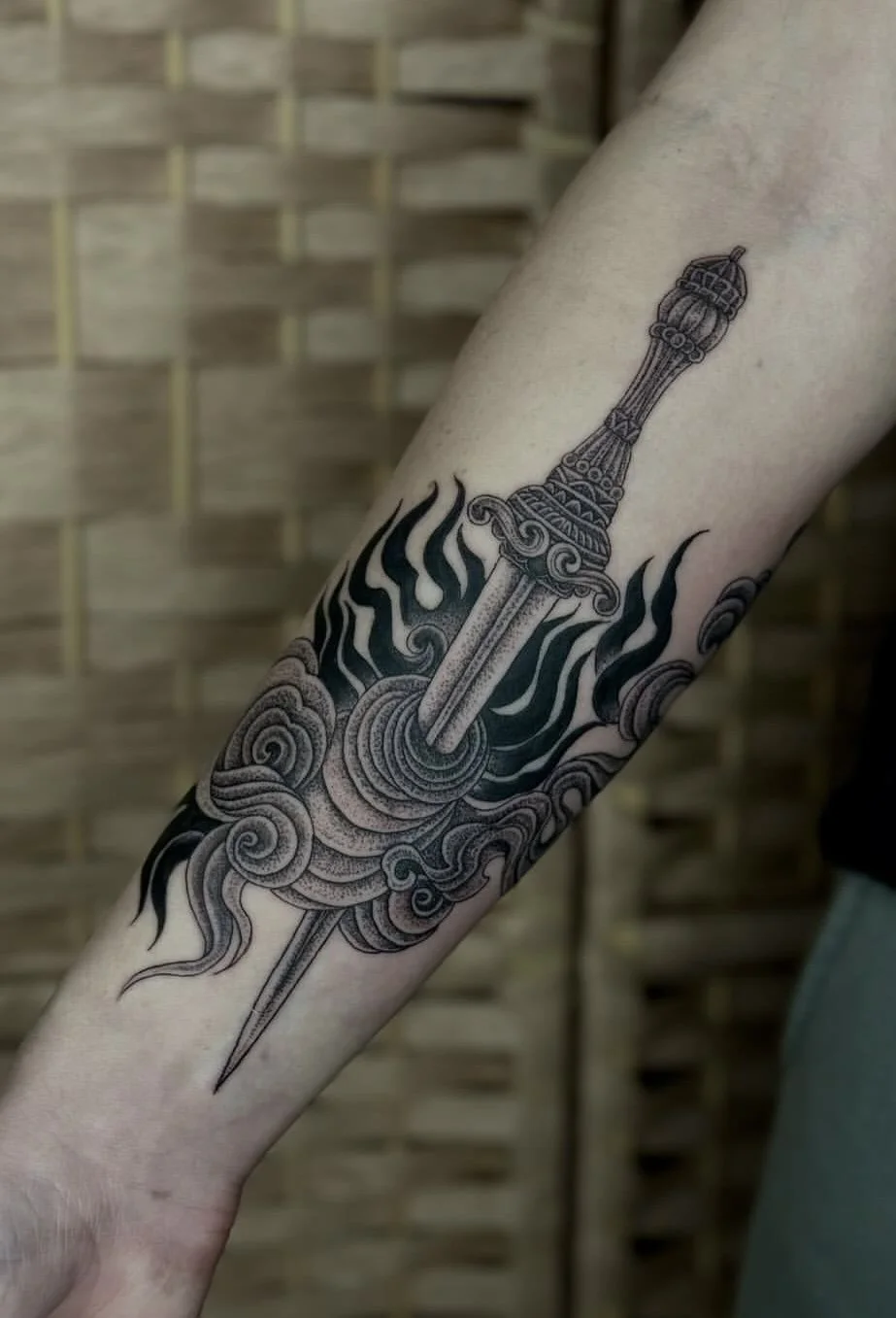 Tattoo of a dagger surrounded by swirling clouds and smoke on a person's forearm.