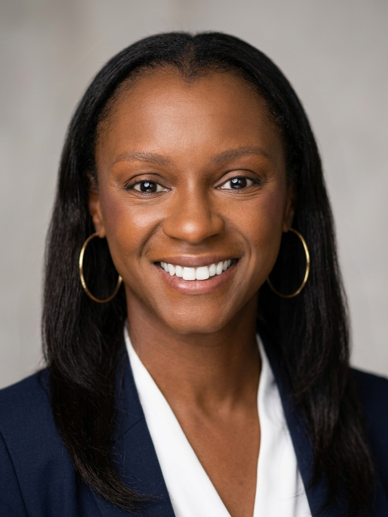 A smiling woman with dark hair wearing hoop earrings, a navy blazer, and a white blouse, posing against a plain background.
