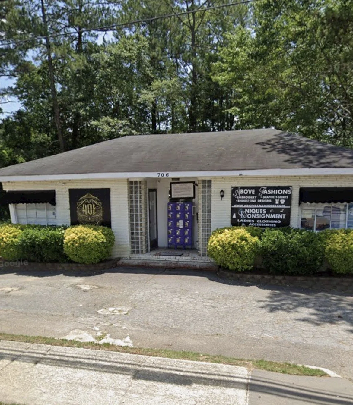 Photo of a small white building with black signs advertising a clothing and accessories store, with green bushes in front and trees in the background.