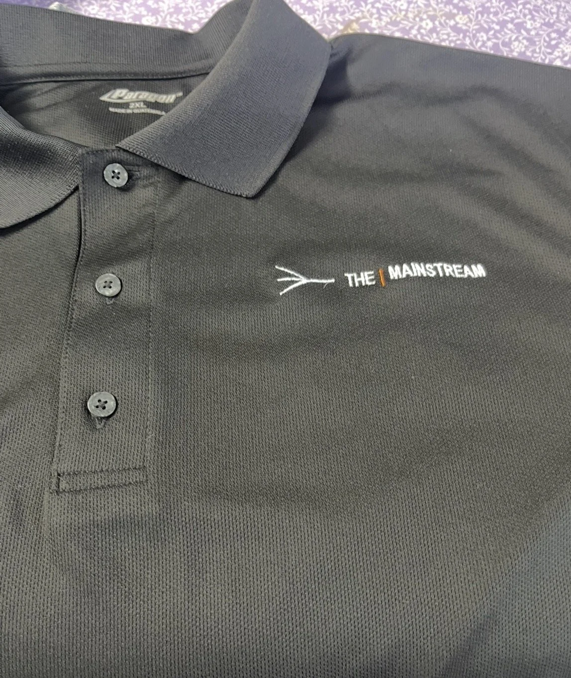 A black polo shirt with a logo and text embroidered on the left chest area. The logo is 'THE MAINSTREAM' with a small orange line separating 'THE' and 'MAINSTREAM'.