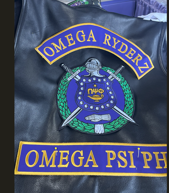 lack leather motorcycle vest with an Omega Rydez top rocker patch, an Omega Psi Phi bottom rocker patch and a shield with swords and Greek letters patch sewn onto the back of the vest.