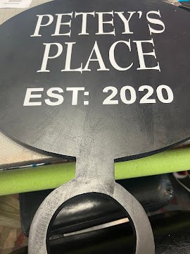 Cutting board with "PETEY'S PLACE EST: 2020" in white vinyl on a black paddle-shape board.