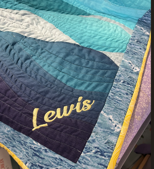 quilted blanket with wave patterns in various shades of blue, white, and purple, with the name 'Lewis' embroidered in yellow on the corner.