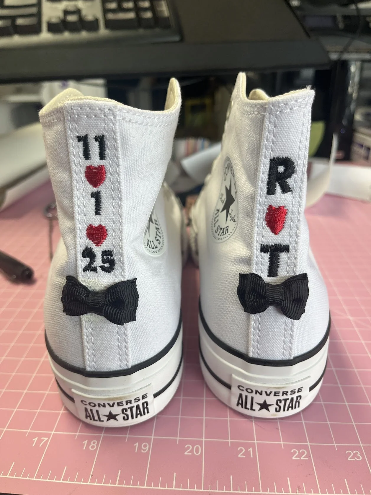 Rear view of white Converse All Star high-top sneakers decorated with embroidered text and symbols 11 ♥ 1 25' on the left shoe and 'R ♥ T' on the right shoe, and embellished with black bows on the back.