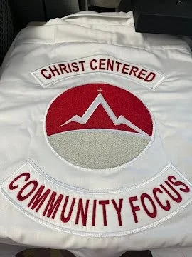 White shirt with a embroidered circular logo of a mountain with a cross at the peak inside a red circle. The lettering says, 'Christ Centered' at the top and 'Community Focus' at the bottom in red letters.