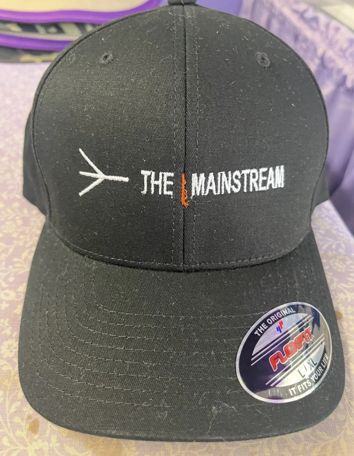 Black baseball cap with embroidered text 'THE MAINSTREAM' with a small orange line separating 'THE' and 'MAINSTREAM'.