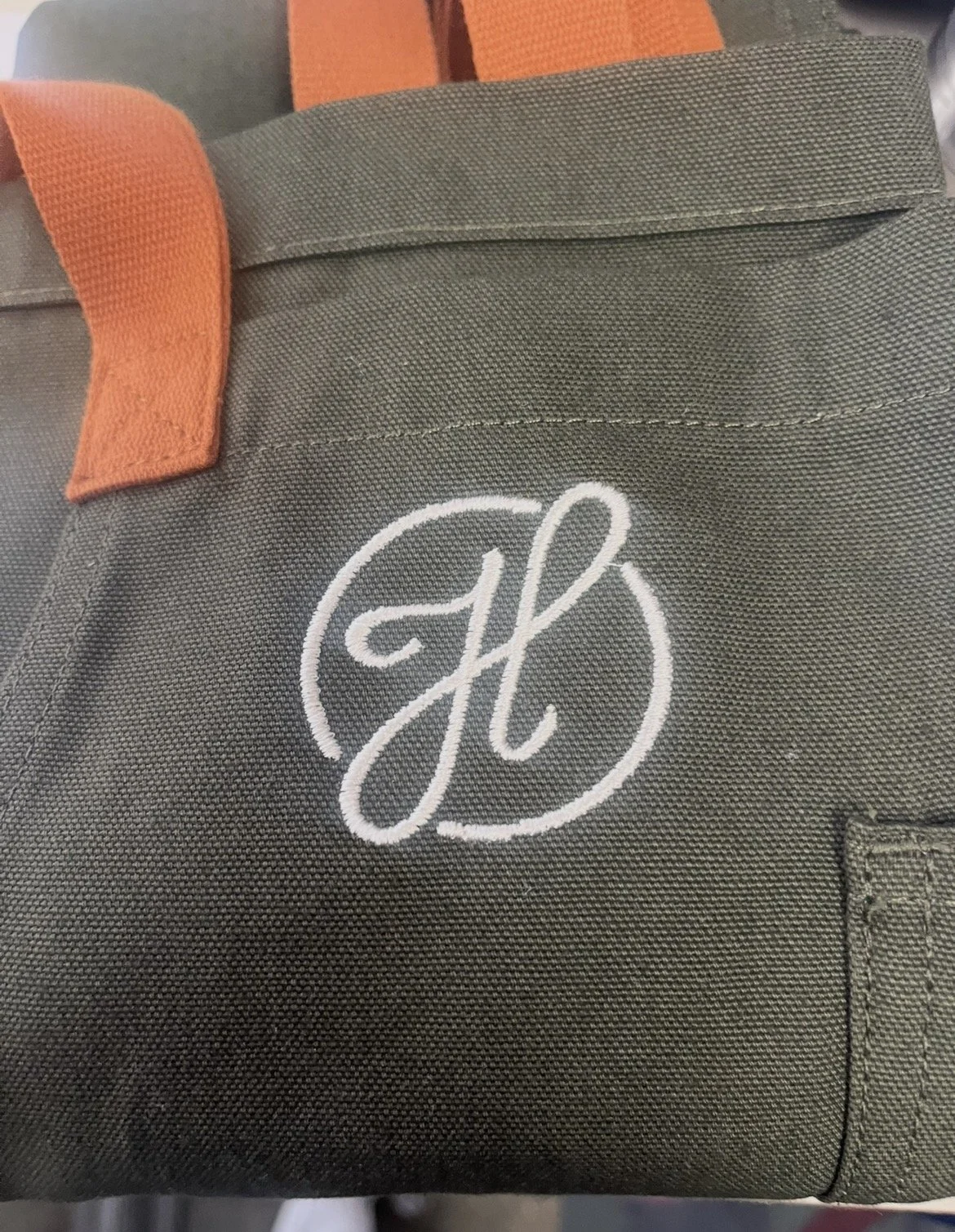 A green fabric apron with leather straps and a cream embroidered monogram 'H' on the front corner.