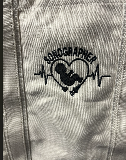 A canvas tote featuring an embroidered logo of a baby in a heart, with a heartbeat line, and the word 'SONOGRAPHER.