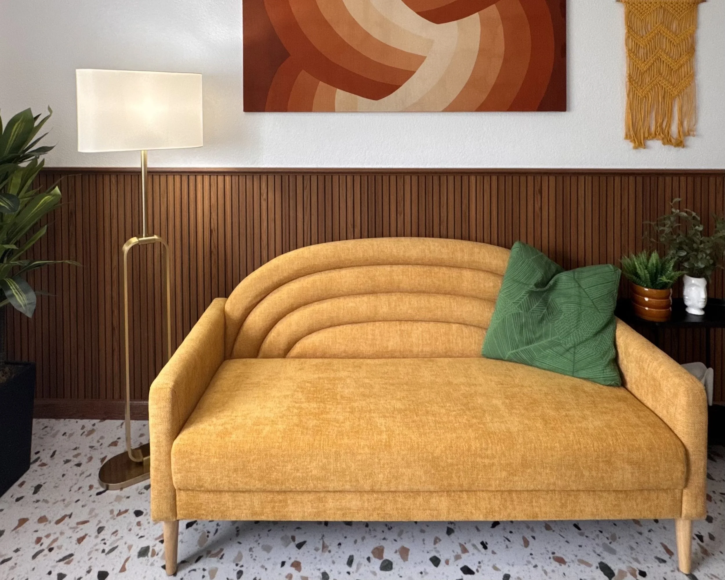 A mid-century modern yellow sofa with curved backrest and wooden legs, decorated with green cushions, in a room with wood-paneled wall, abstract orange wall art, and a tall floor lamp, beside a potted plant and a small white sculpture.