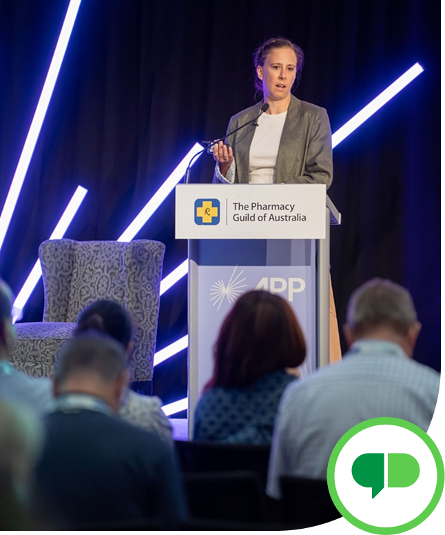 A woman standing at a podium labeled 'The Pharmacy Guild of Australia' speaking to an audience at a conference, with a decorative black curtain and blue neon light strips in the background.
