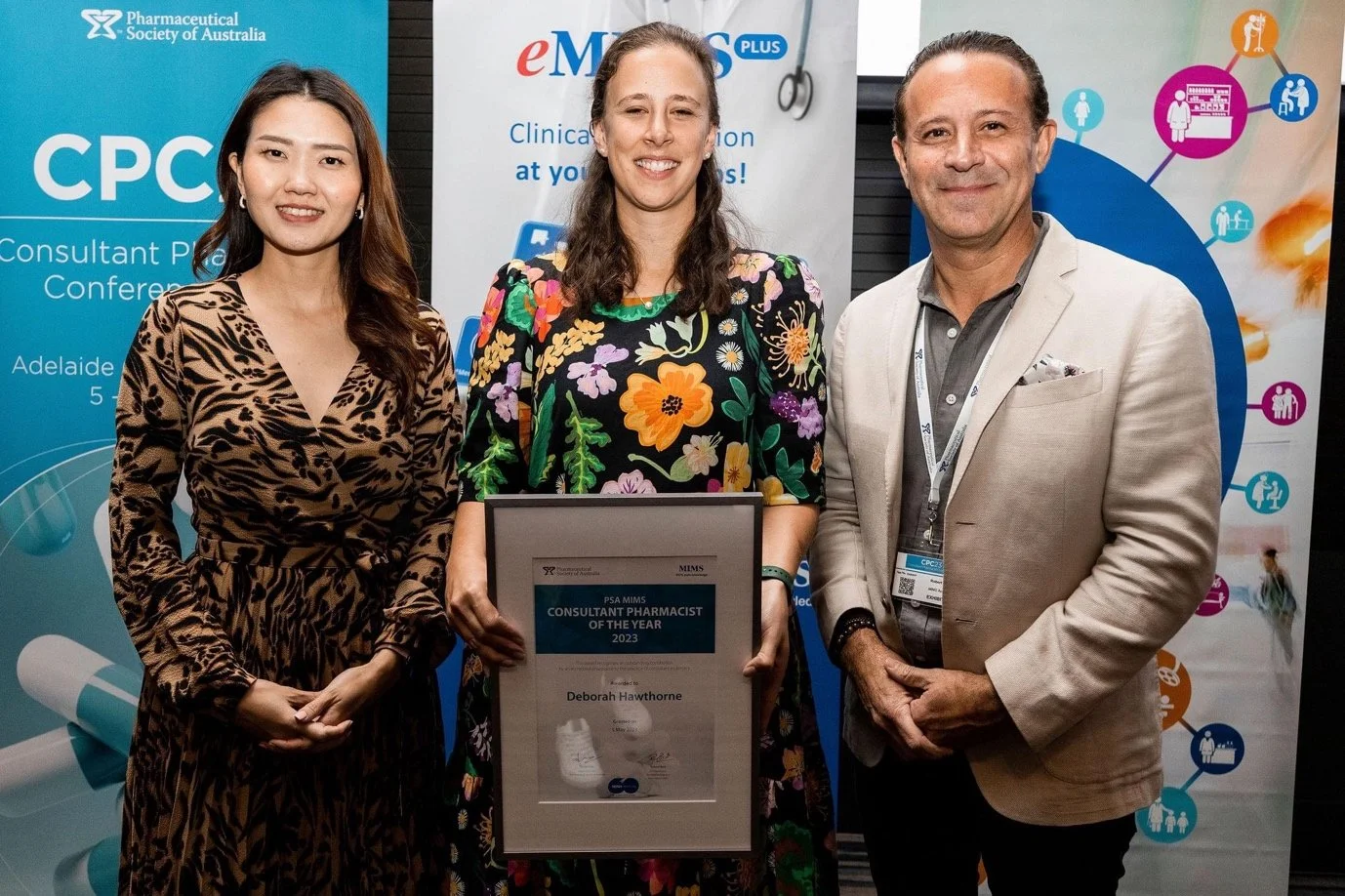 Three people standing together, two women and one man, at an awards event. The woman in the middle is holding a framed certificate that reads "Consultant Pharmacist of the Year 2023." They are in front of banners related to a pharmaceutical conference.