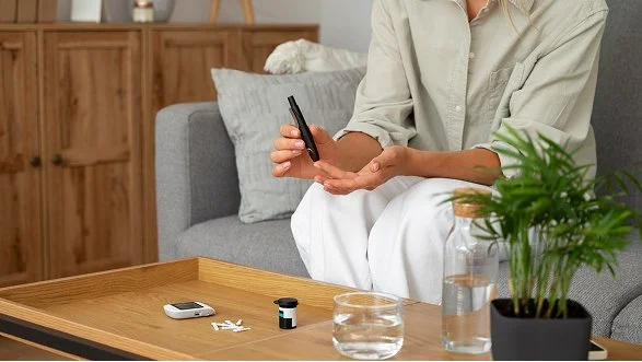 A person in beige shirt and white pants sitting on a gray couch, holding a bottle of medicine or supplement. On a wooden coffee table in front of them are a pill bottle, some pills, a glass of water, and a green potted plant.