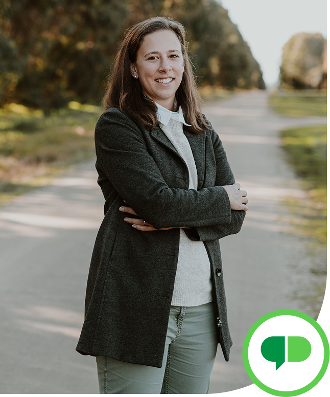 A woman standing outdoors on a pathway, smiling with arms crossed, wearing a dark coat, white shirt, and gray pants, with trees and greenery in the background.
