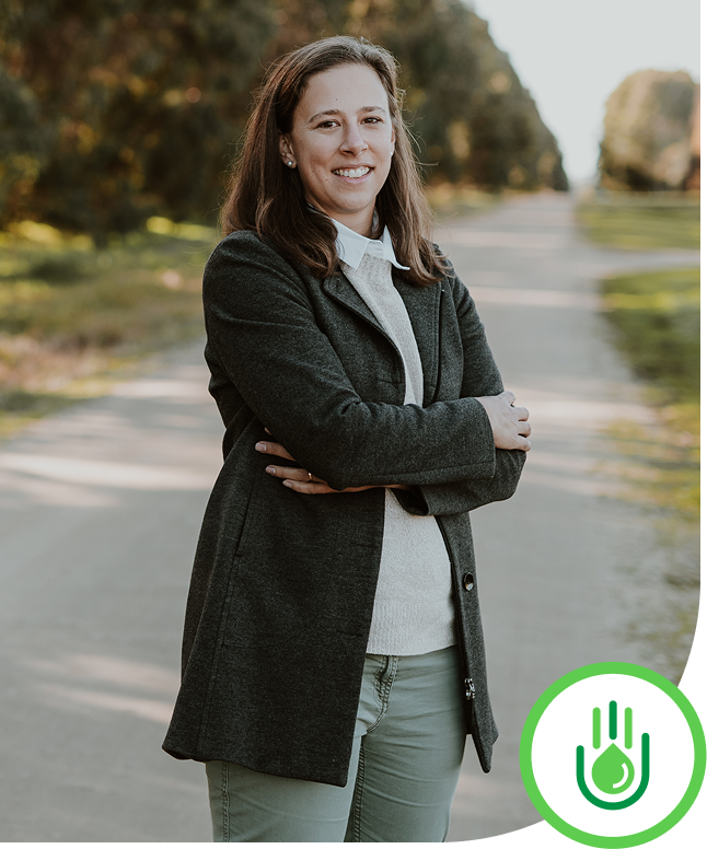 A woman with brown hair and a light skin tone standing outdoors on a rural road with trees on both sides, arms crossed, smiling, wearing a gray coat over a light sweater and light-colored pants.